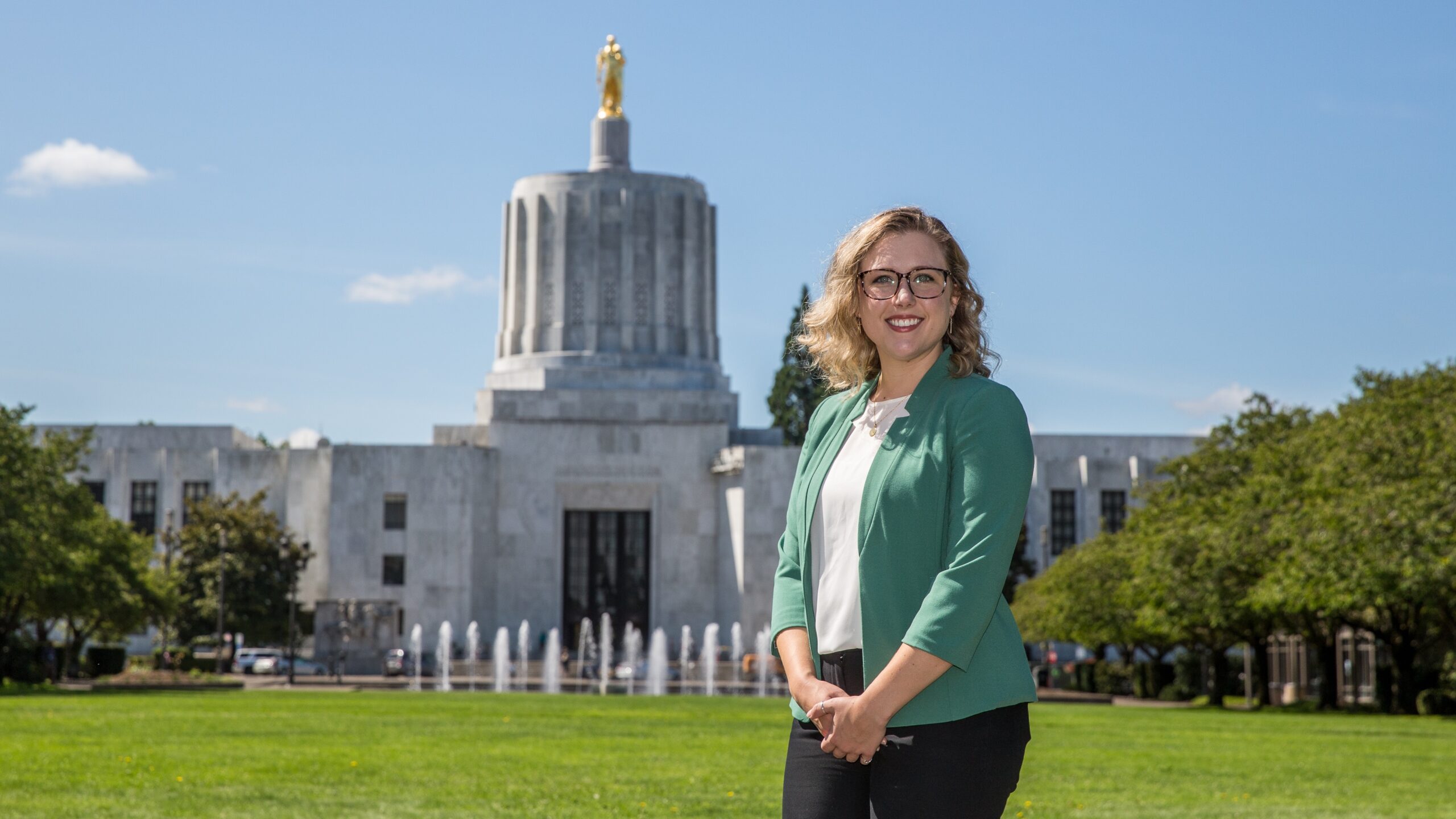 A woman with glasses poses in front of the Oregon State Capitol, highlighting the building's iconic architecture.