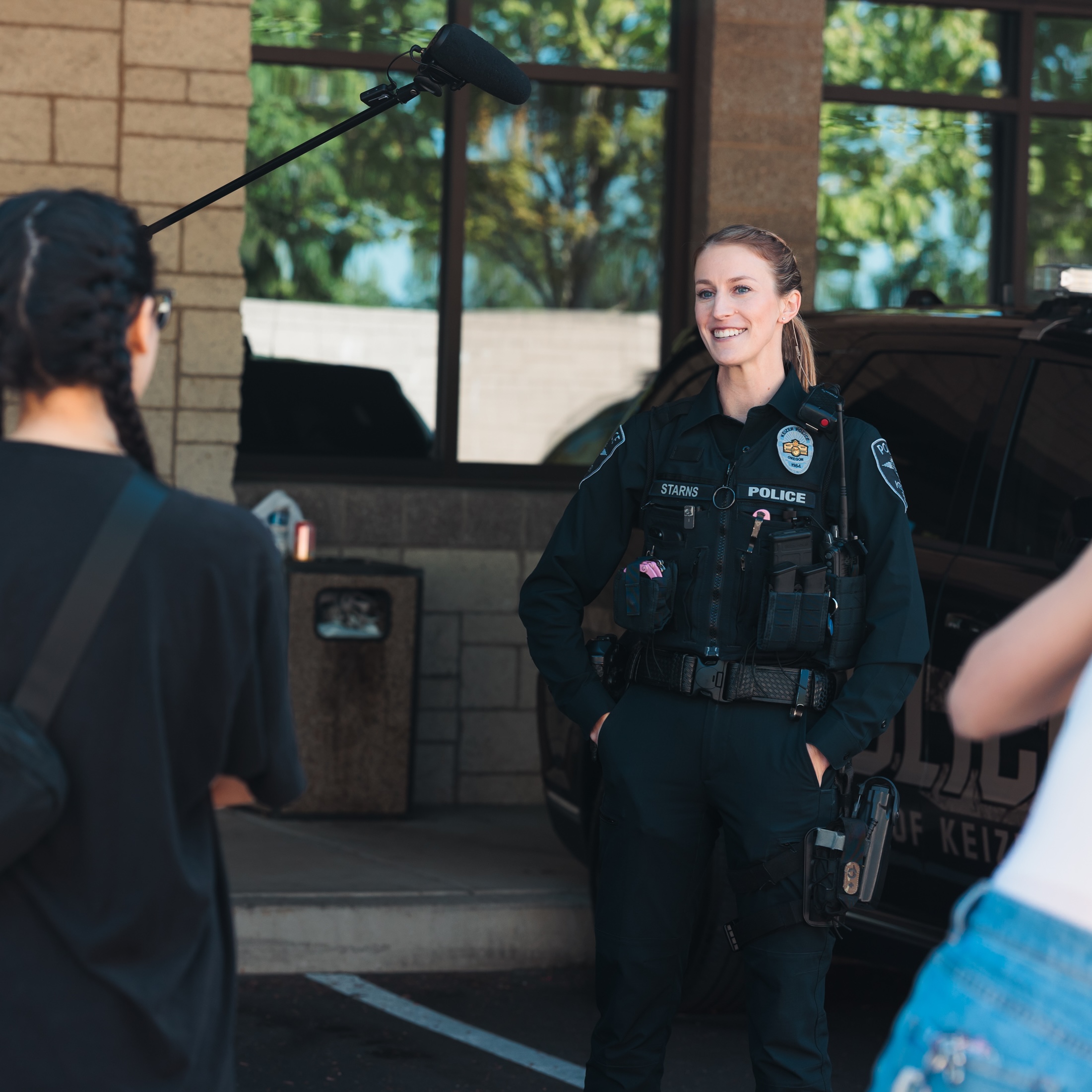 A woman dressed in a police uniform stands beside a car for a media shoot as a student interviews her.