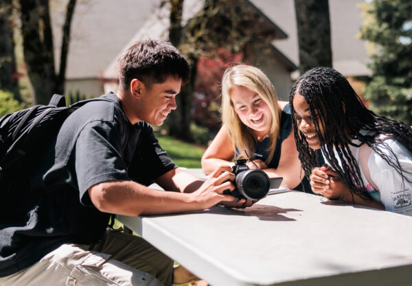 Two students and one professor gather at a picnic table, one holding a camera, discussing its features.