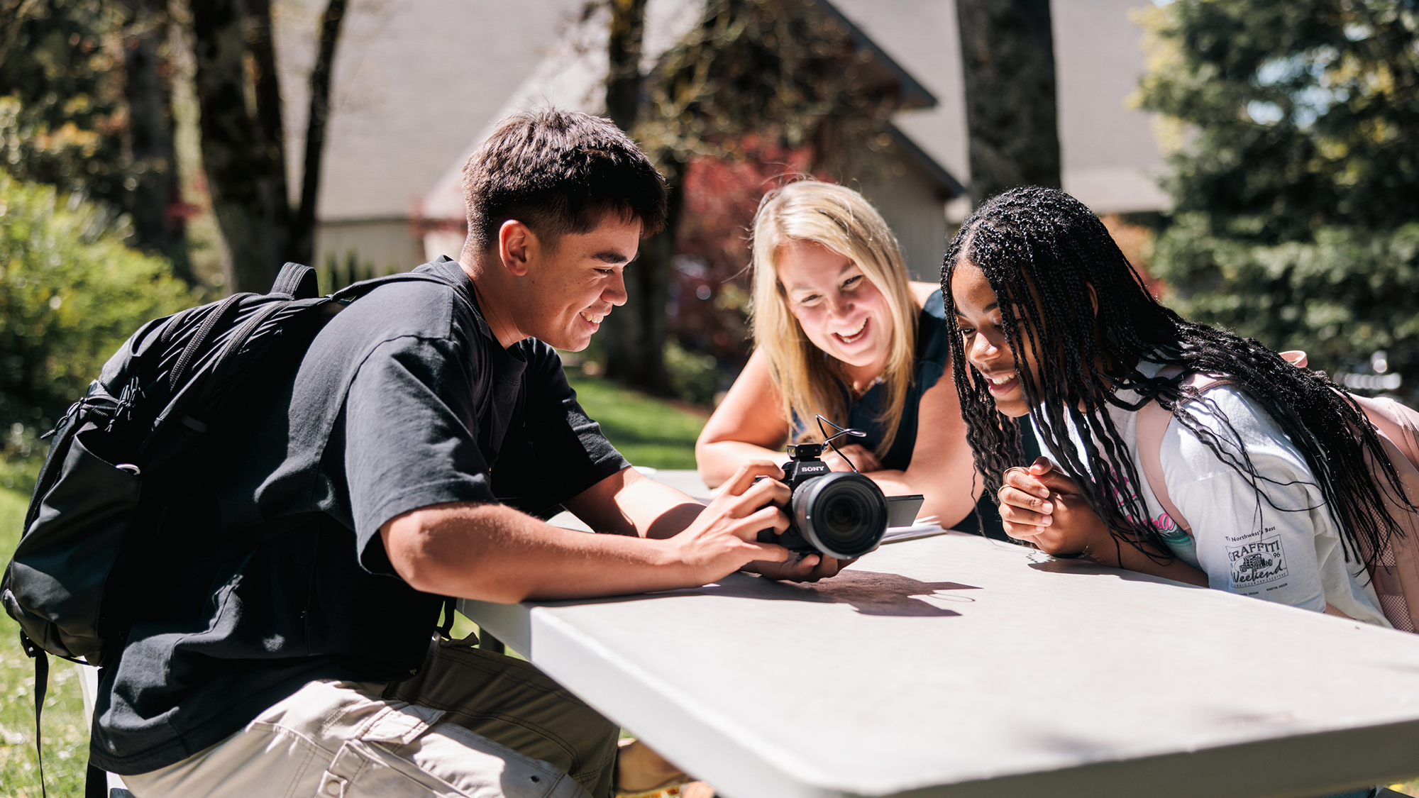 Two students and one professor gather at a picnic table, one holding a camera, discussing its features.