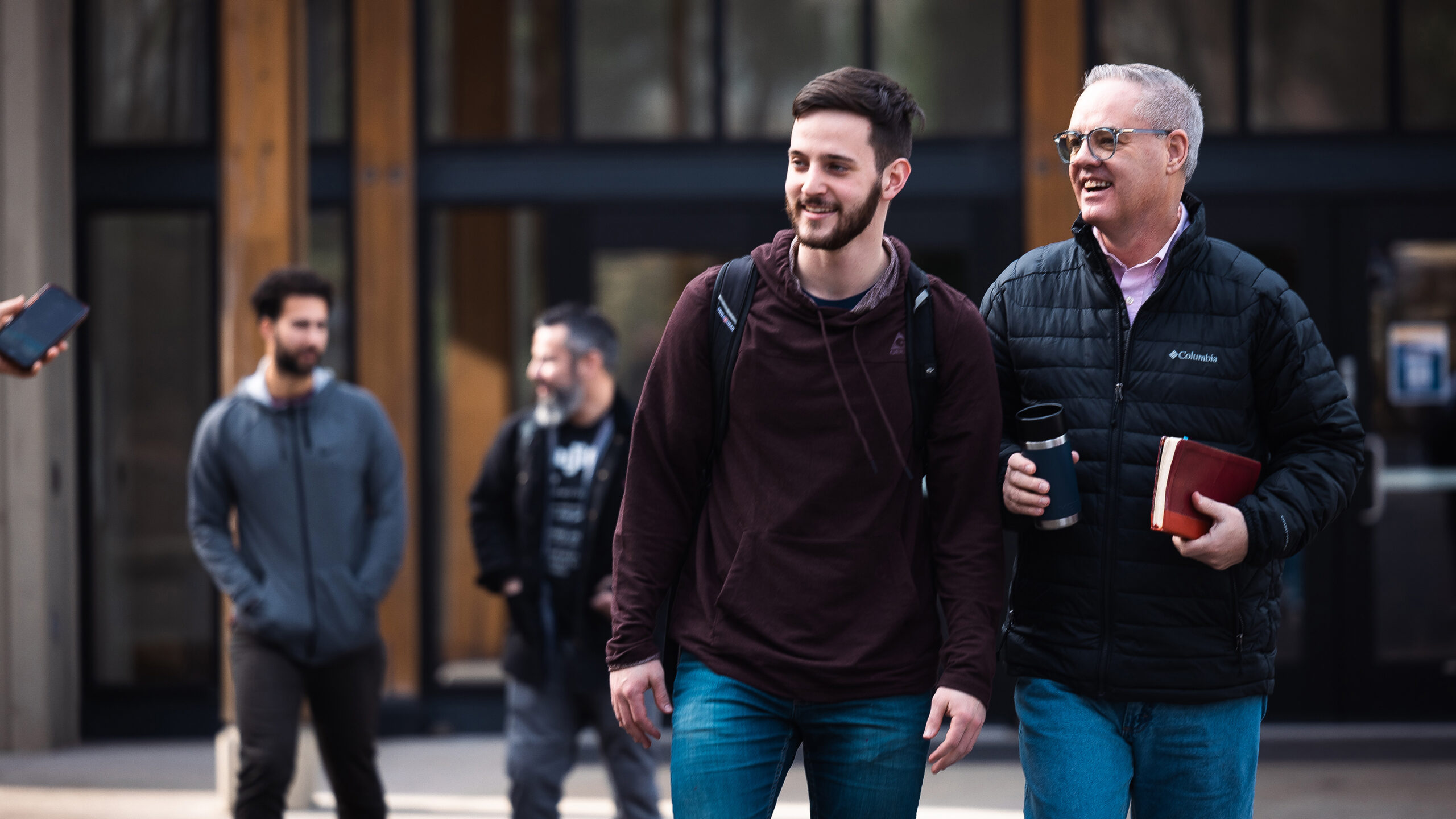 A younger man with a backpack and an older man holding a coffee cup and a book walk together outside, smiling. Two other men in casual clothing are in the background, also smiling while walking out of chapel.