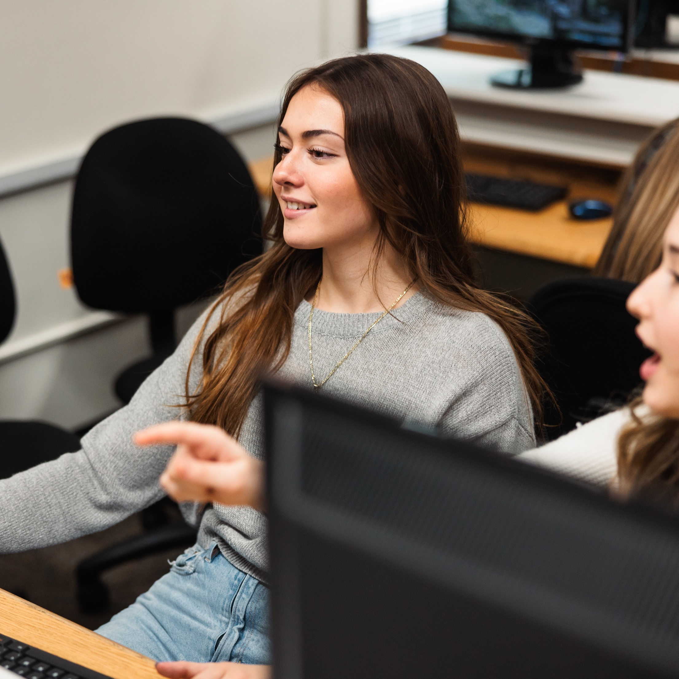 Two women engaged in discussion while working at a computer in a computer lab.
