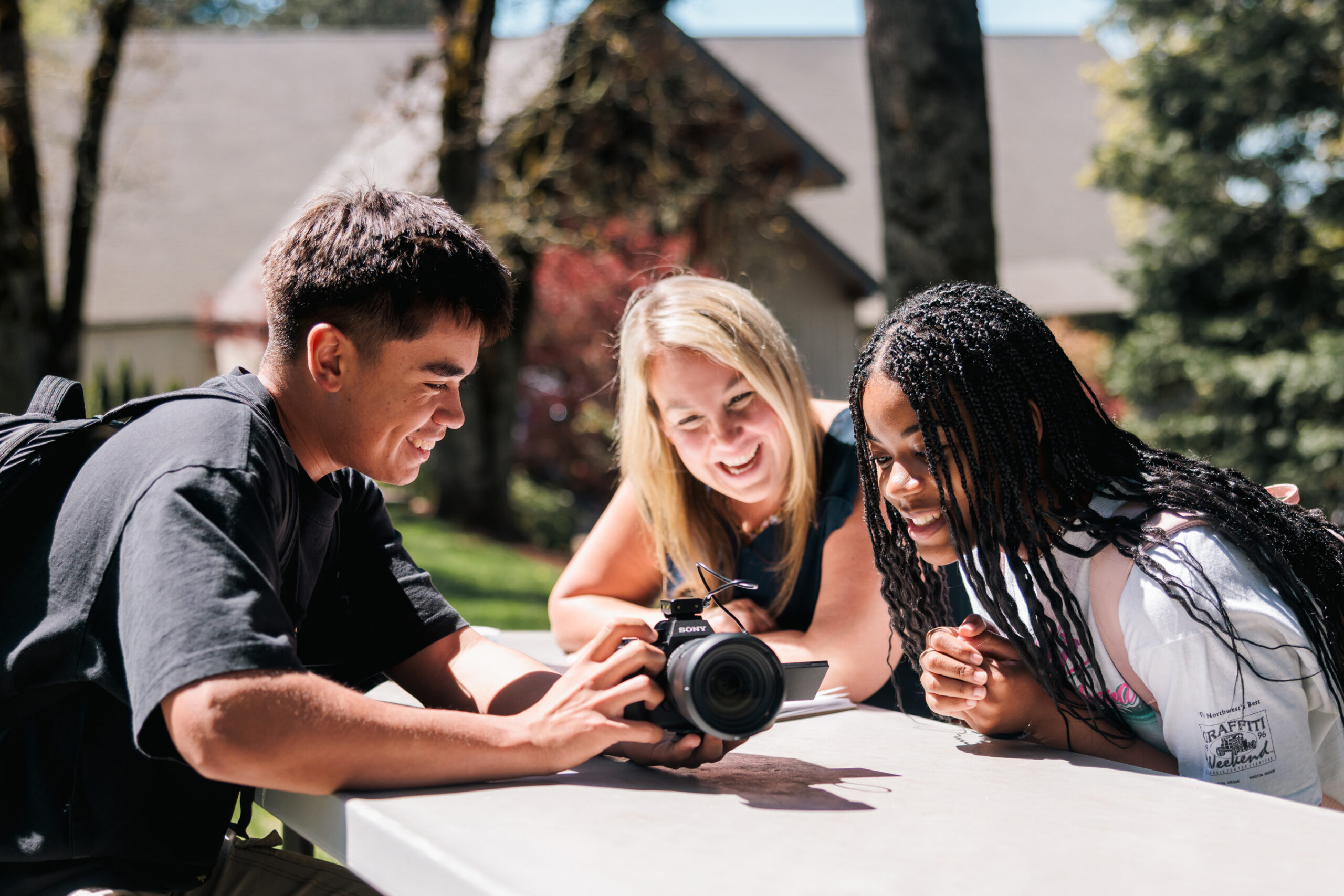 Two students are seated at an outdoor table with their professor, holding a camera and laughing as they discuss photography.