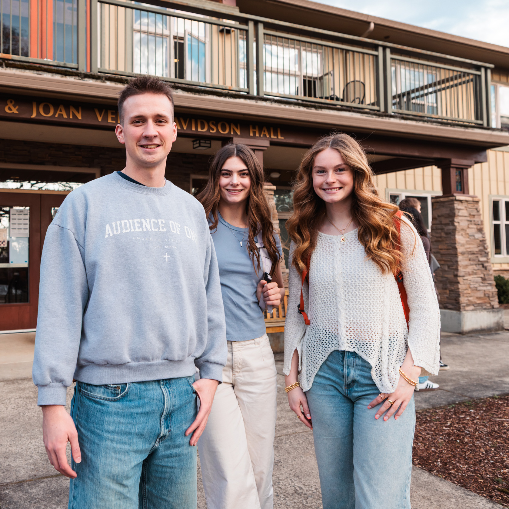 Three college students--a man and two women--smile at the camera in front of Davidson Hall, a cabin-style residence hall.