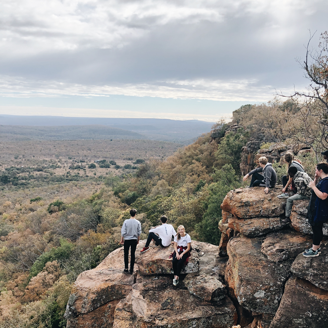 A group of people sitting on a cliff's edge, enjoying a stunning view of a valley in Africa.
