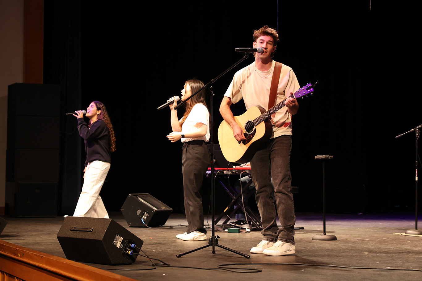 Three musicians perform on stage during chapel worship time: one plays guitar and sings, while two others sing into microphones. A keyboard and speakers are also visible.