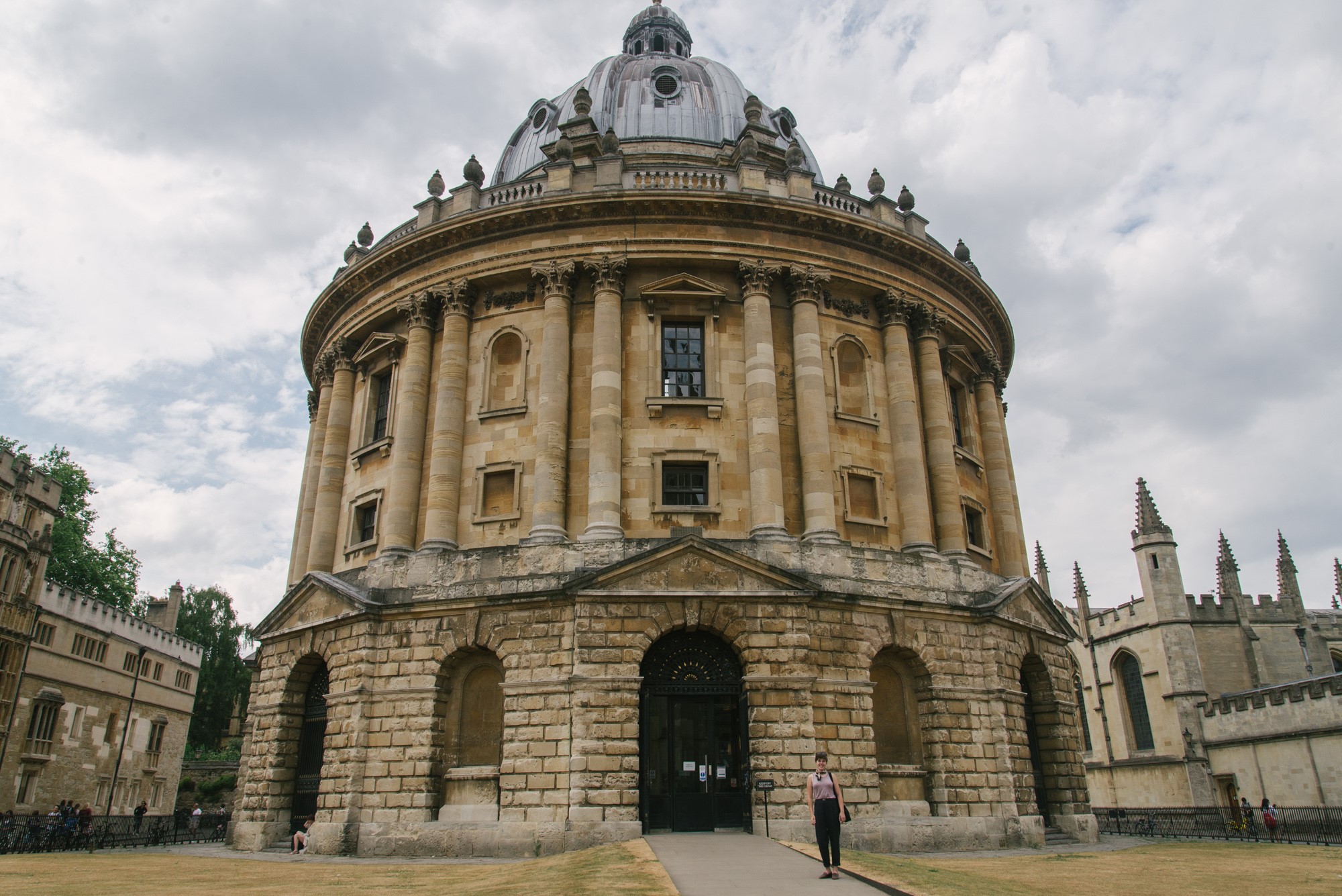A person stands in front of the Radcliffe Camera, an iconic round building with tall columns and a domed roof, located in Oxford, England. The sky is overcast, and other historic buildings are visible in the background.