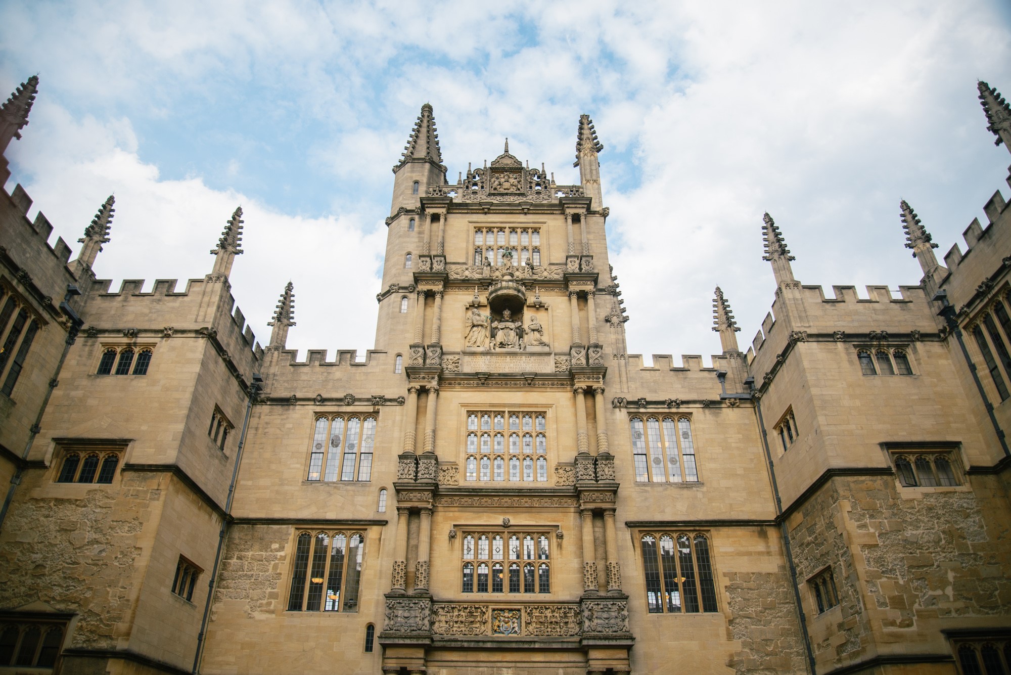 A grand, symmetrical Gothic-style building at the University of Oxford, featuring ornate stone carvings, large mullioned windows, and tall spires along the roofline. The sky above is partly cloudy, and the building’s central tower displays statues and decorative elements.