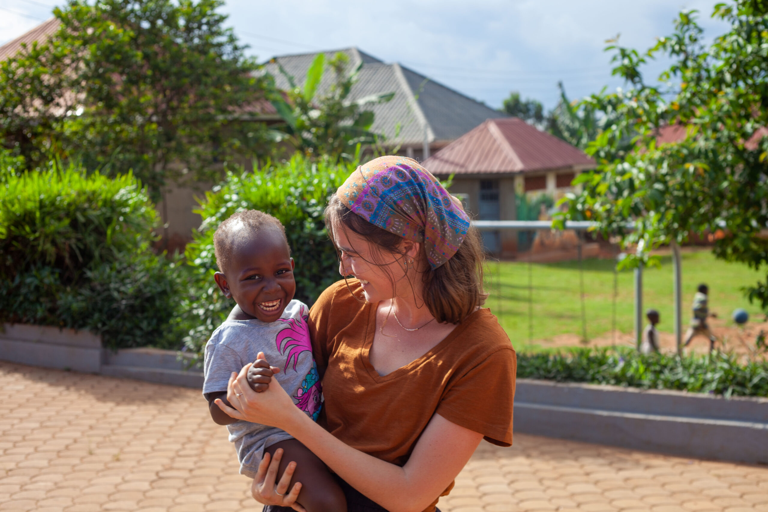 A woman holding a smiling young child outdoors in Uganda, with houses, greenery, and a paved walkway in the background.