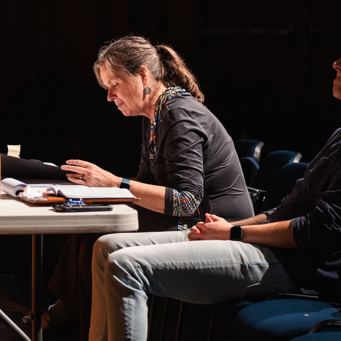 A man and woman seated at a table; Tamara McGinnis reviews notes while observing a theatre practice session.