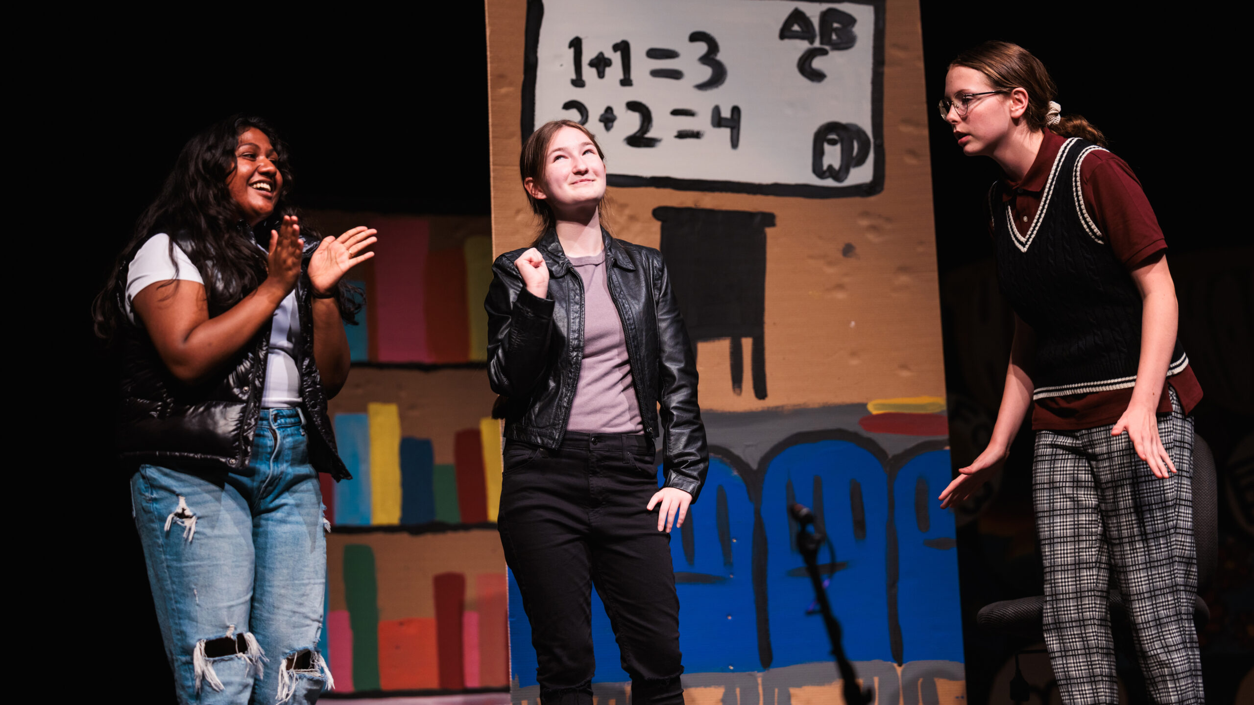 Three high school girls on stage at Theatre Camp, holding a sign that reads "Math," engaging the audience with their performance.