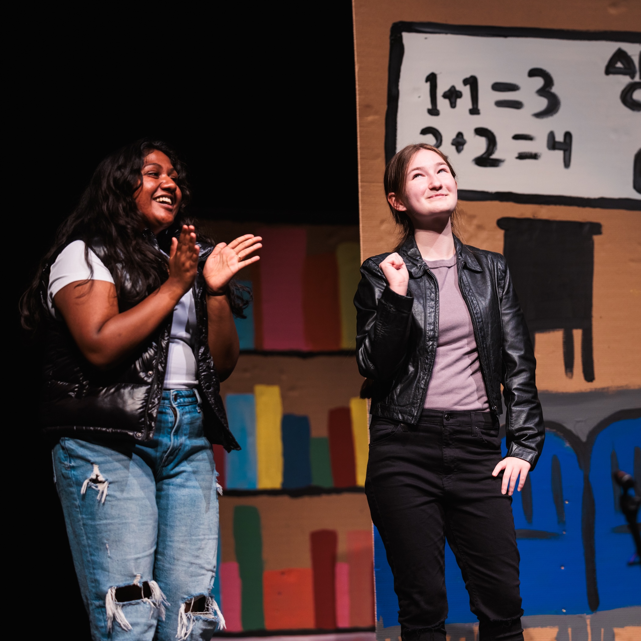 Two high school girls on stage at Theatre Camp, holding a sign that reads "Math," engaging the audience with their performance.