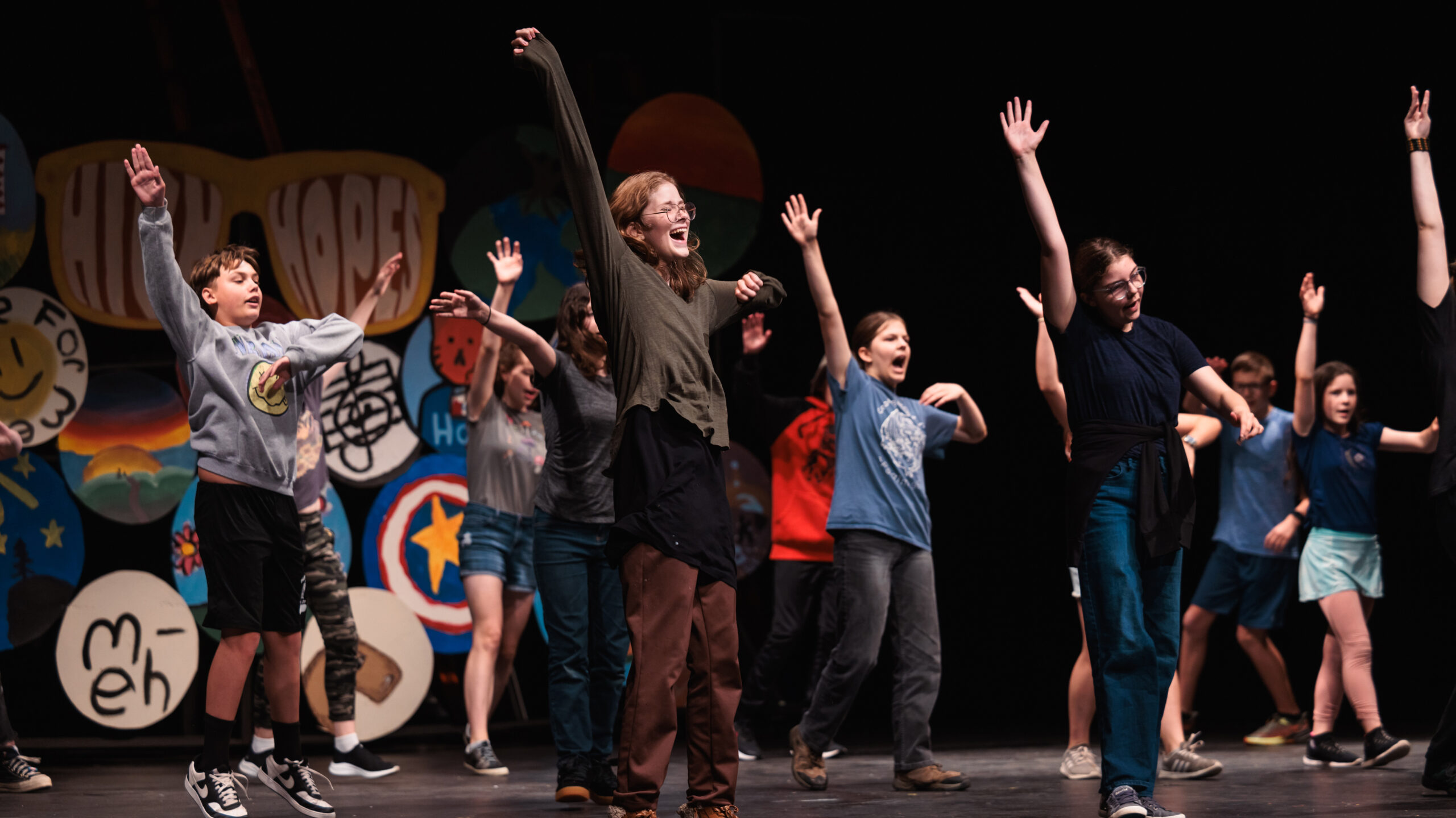 A group of young performers on stage, arms raised, showcasing their song and dance routine during theatre camp.