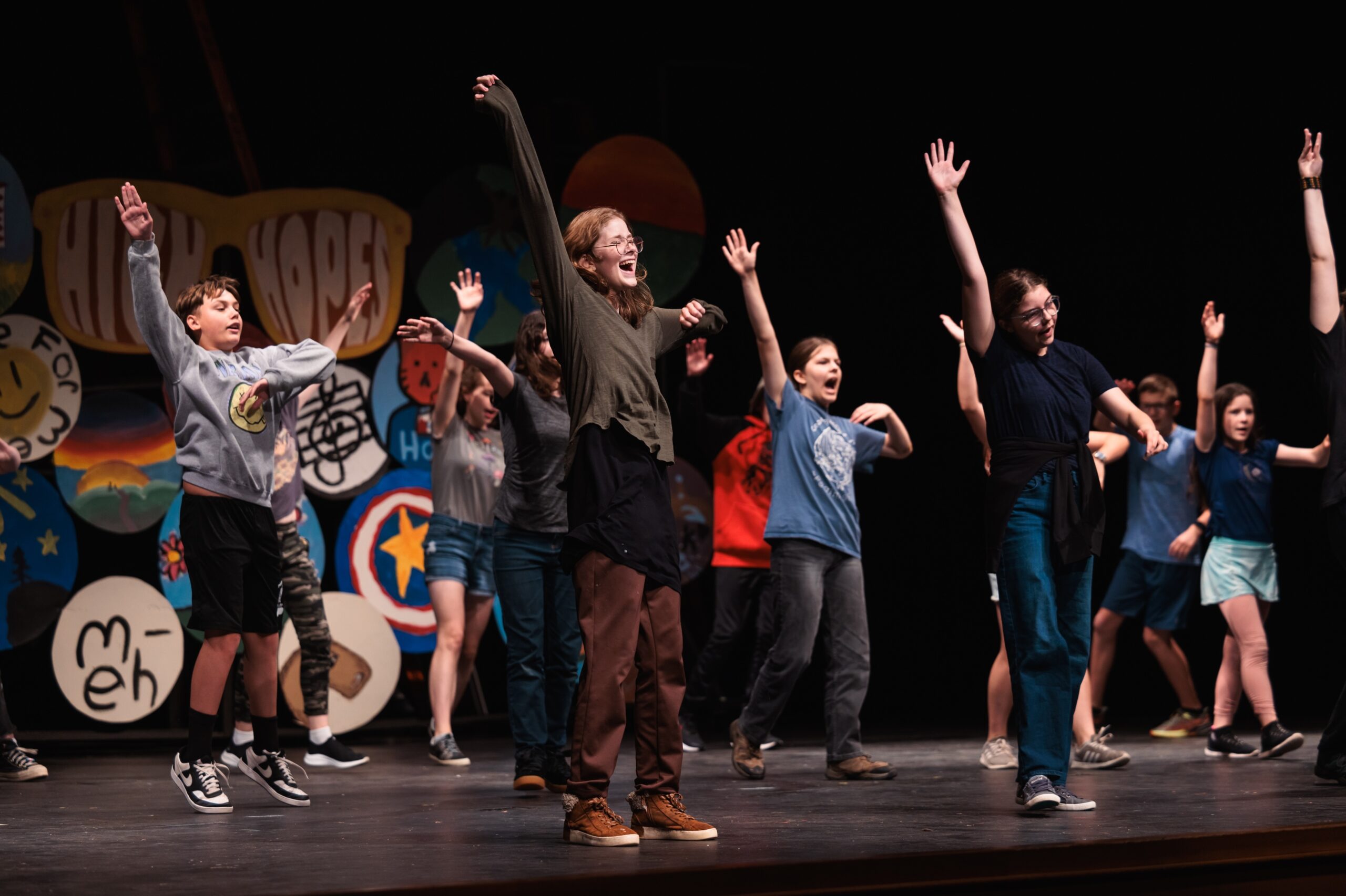 A group of young performers on stage, arms raised, showcasing their song and dance routine during theatre camp.