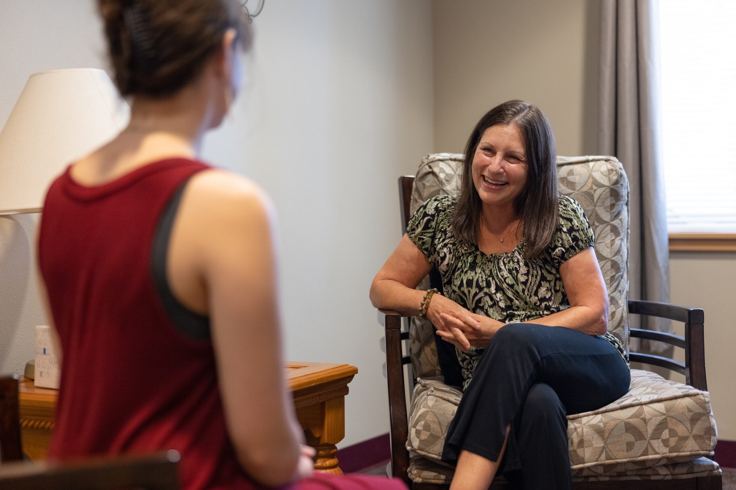 A woman in a floral top sits in a cushioned chair, smiling and talking to another person in a red top who is facing away. They are in a well-lit room with a lamp and window in the background.