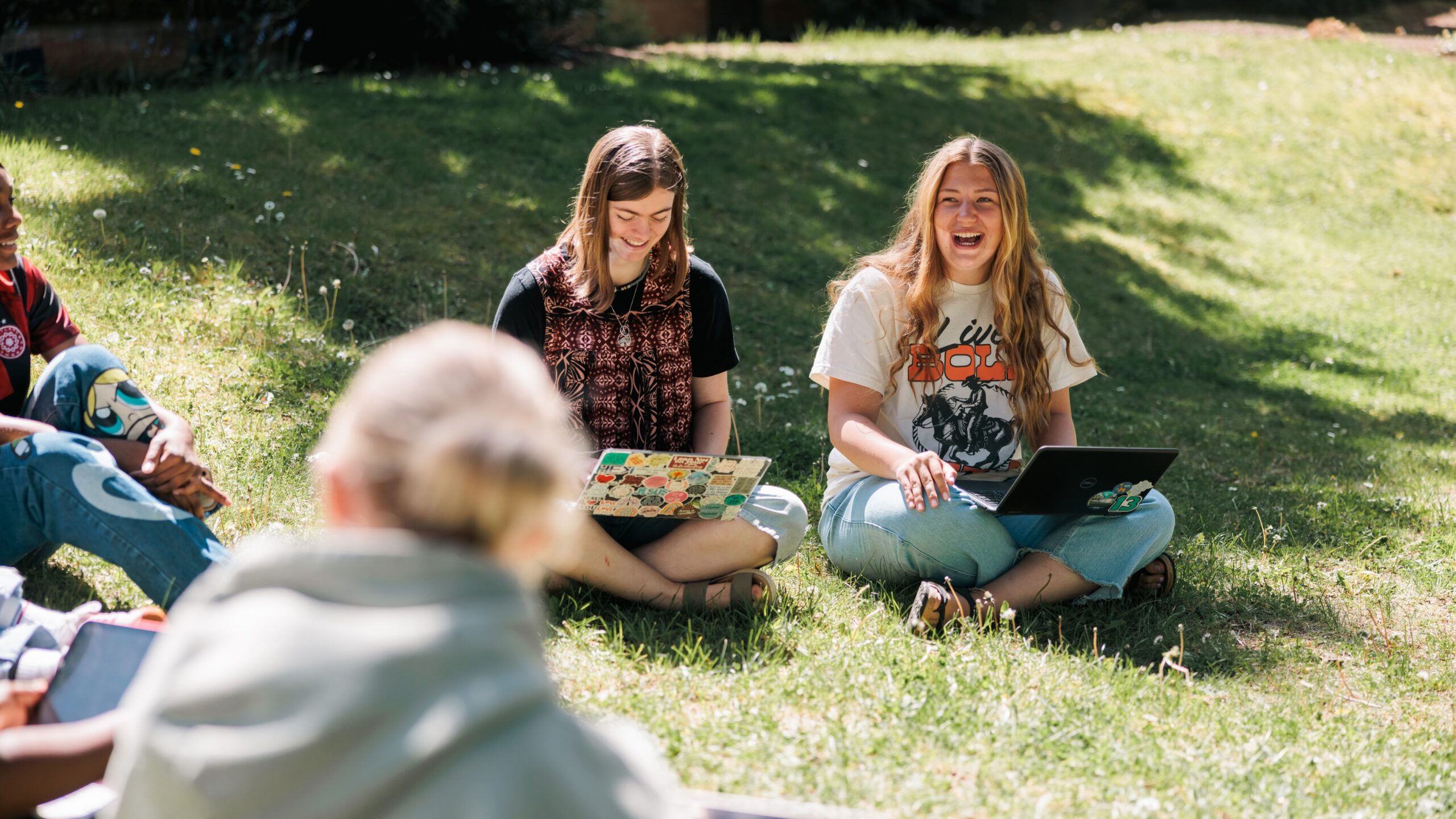 Three young women sit on the grass, each using a laptop, engaged in conversation and collaboration.