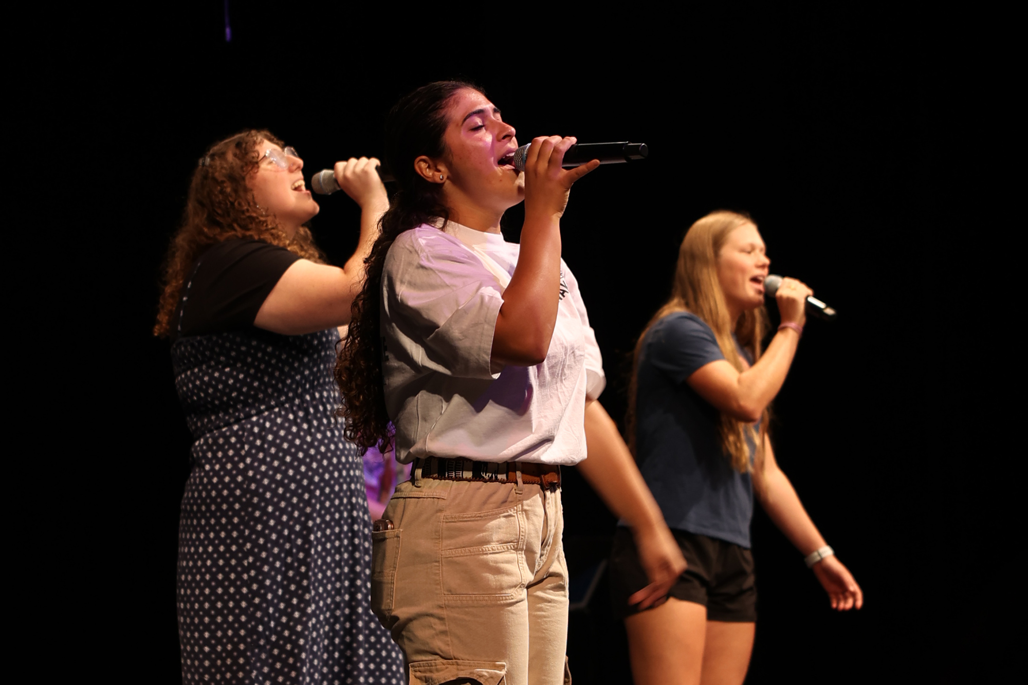 Three young women singing on stage with microphones during the International Student Chapel.