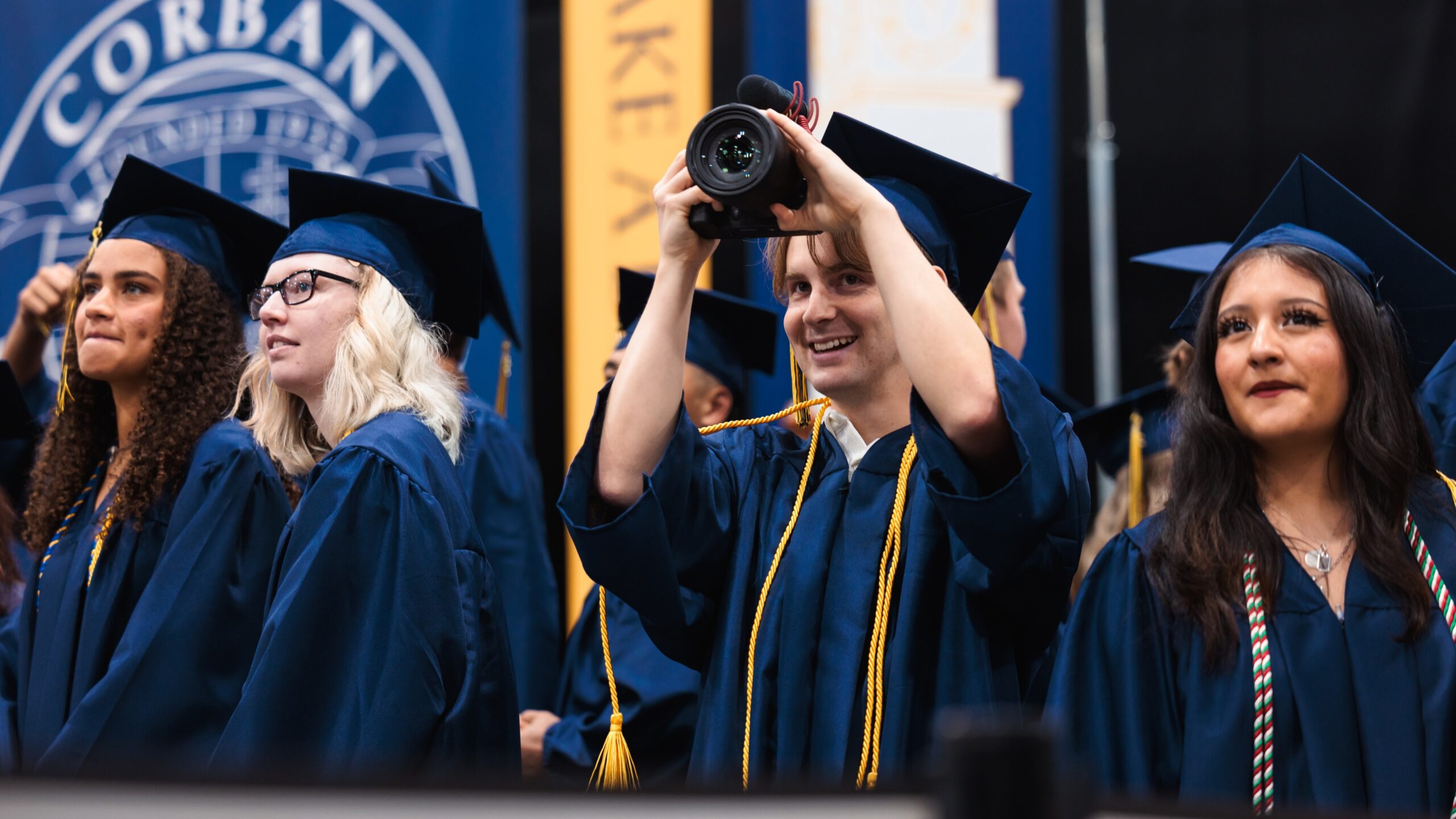 Graduates celebrate at commencement, while one captures the moment on video.