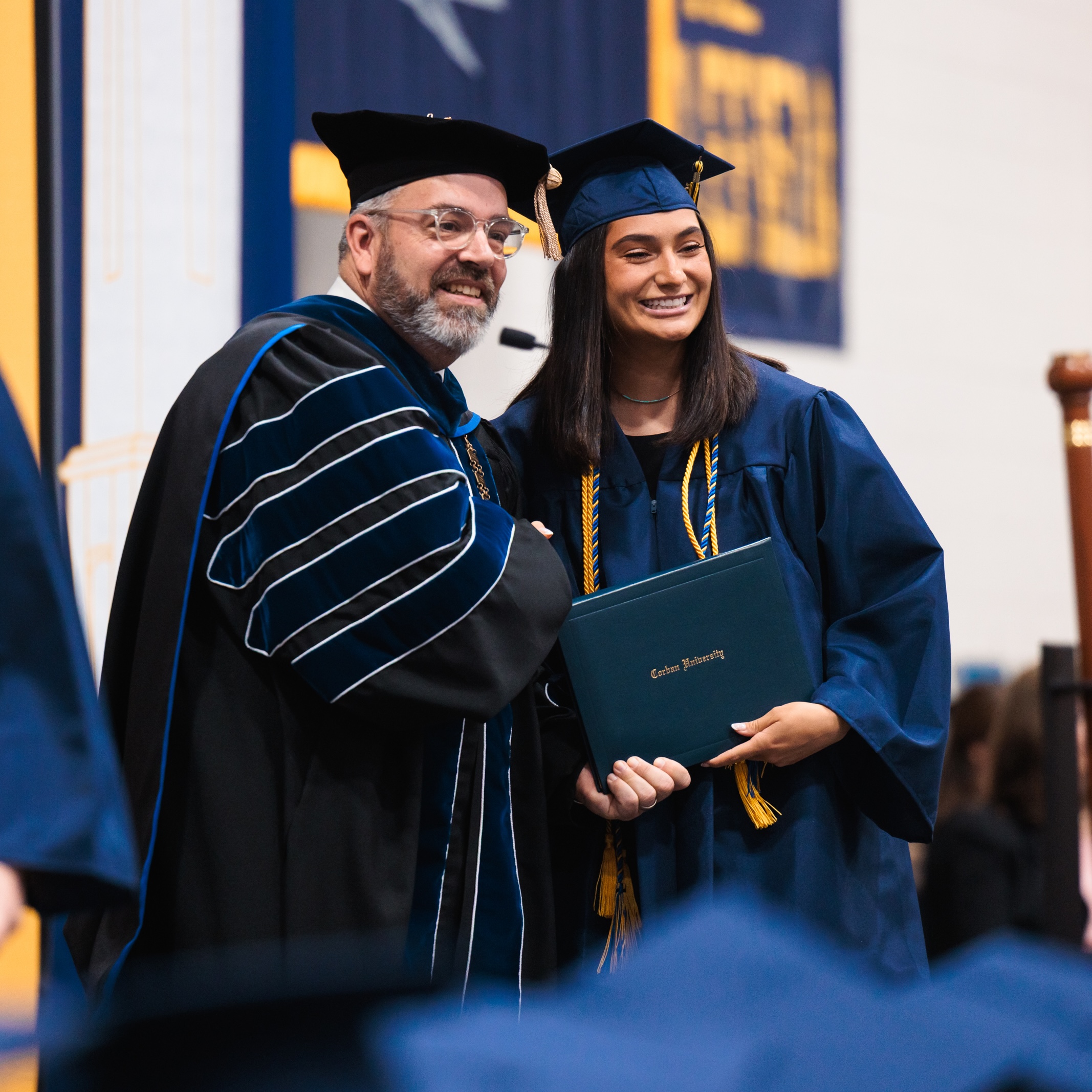 Corban's president shakes hands with a female graduate in their graduation robes at commencement.