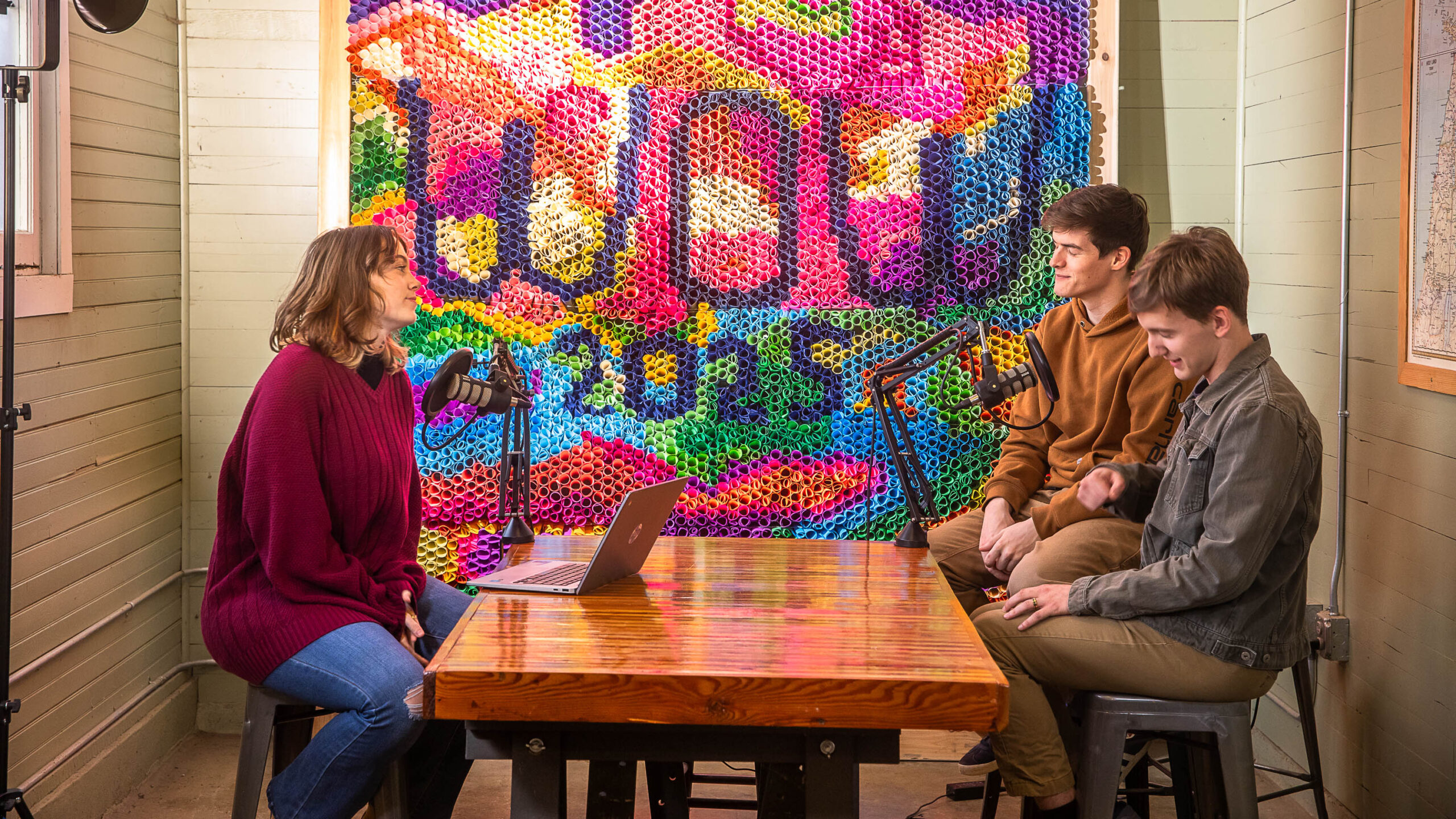 Three students sit in front of a brightly colored backdrop that spells WOW and record a podcast together.