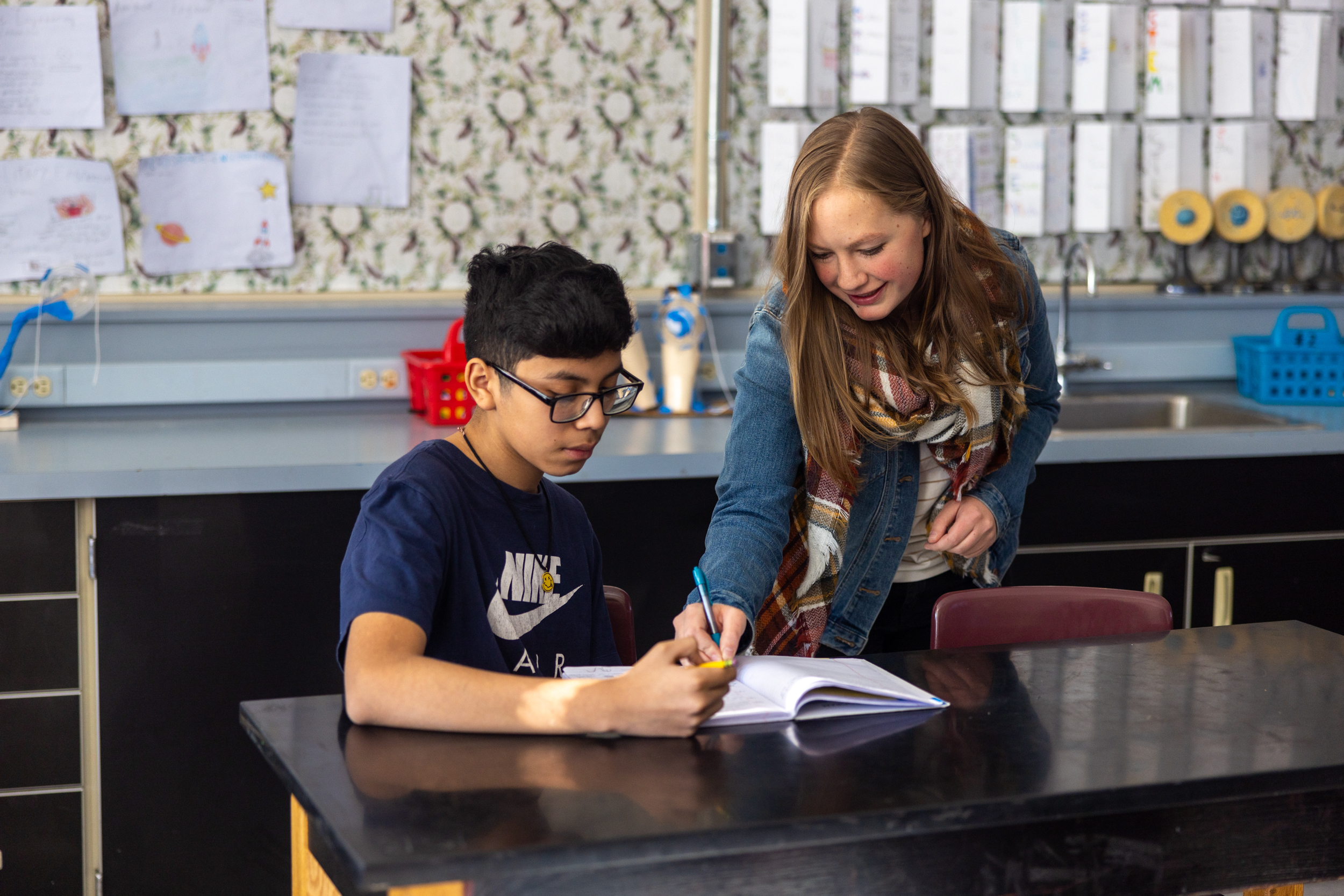 In a classroom, a woman guides a boy as he works on his homework, promoting educational engagement.