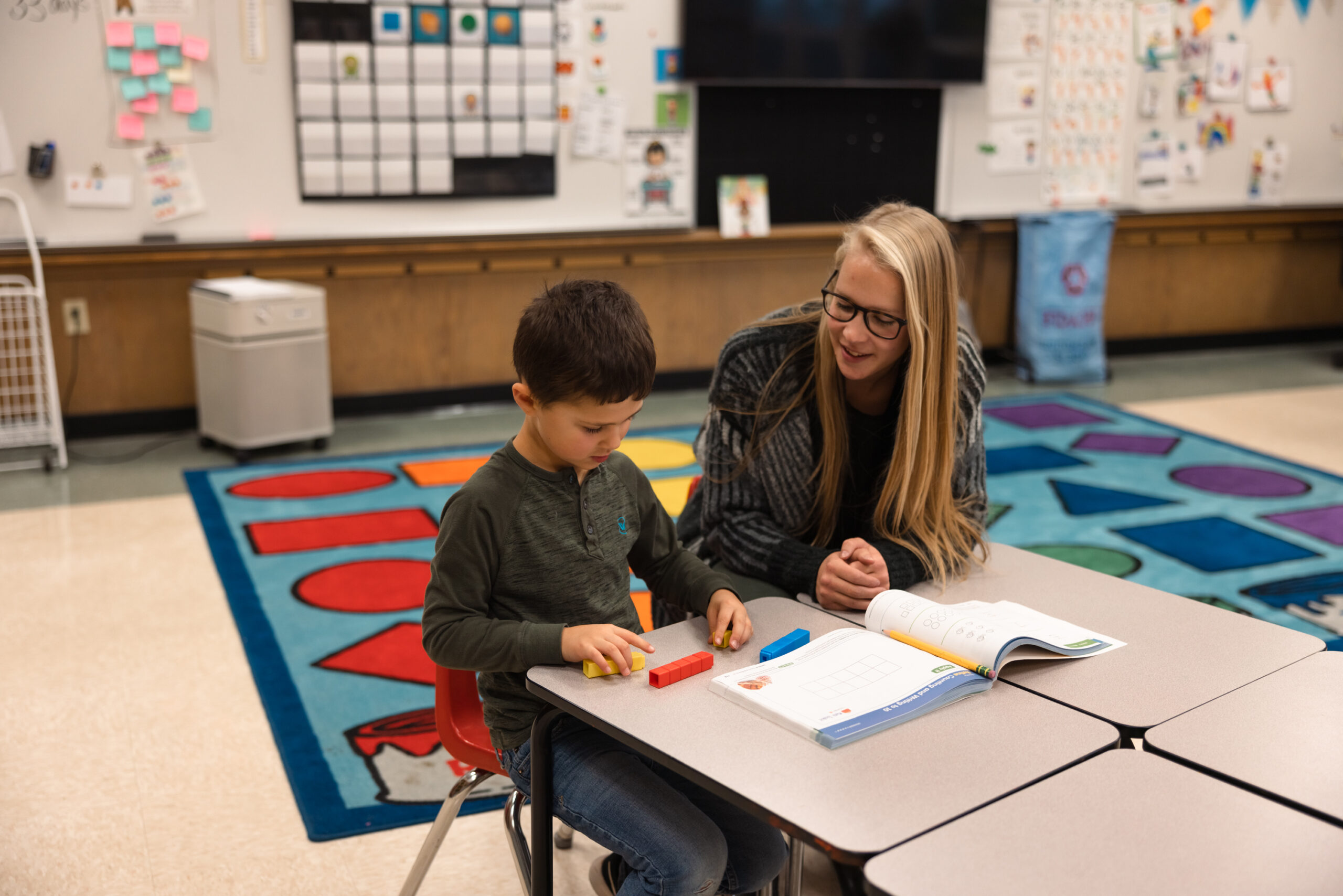 A woman and a boy are seated at a table in a classroom, engaged in an activity together.