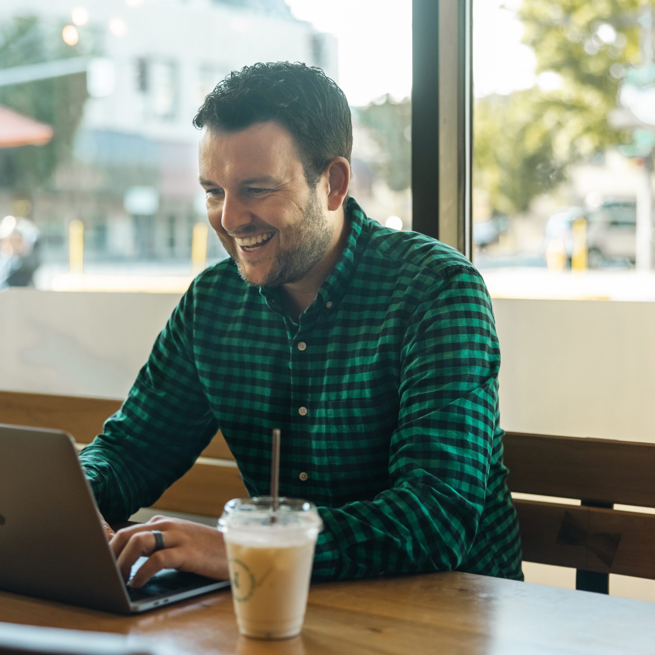 A man in a green checkered shirt is sitting at a wooden table in a café, smiling at his laptop. A cup of iced coffee is on the table. Sunlight filters through large windows behind him, with a street scene outside.