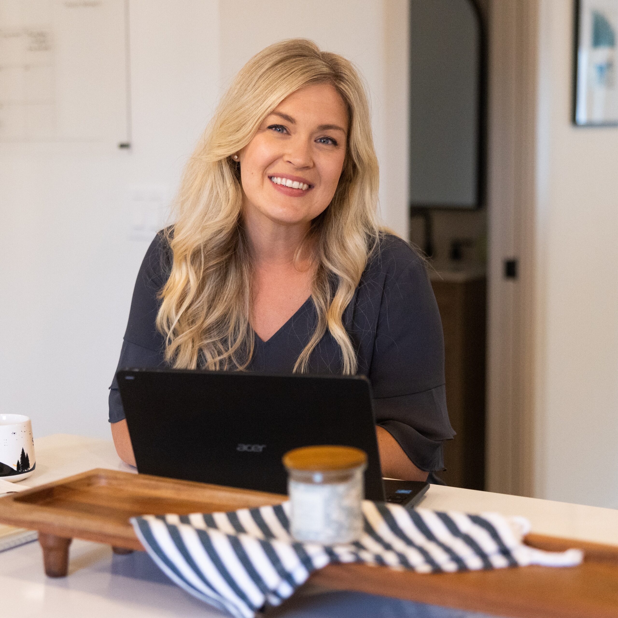 A woman working on a laptop at a table, smiling in a cozy indoor setting.