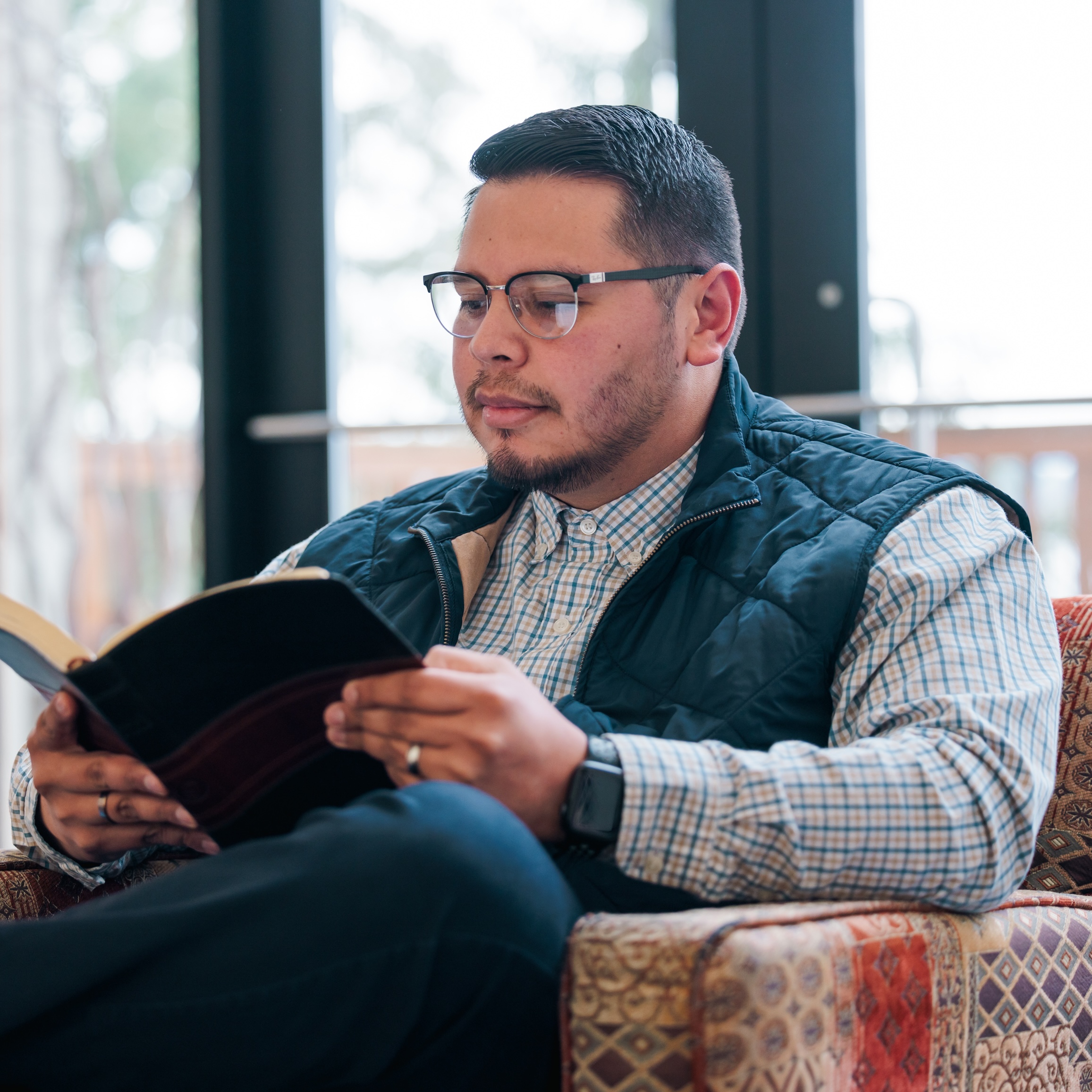 A man in glasses and a vest is intently reading the Bible, seated comfortably in a cozy environment.