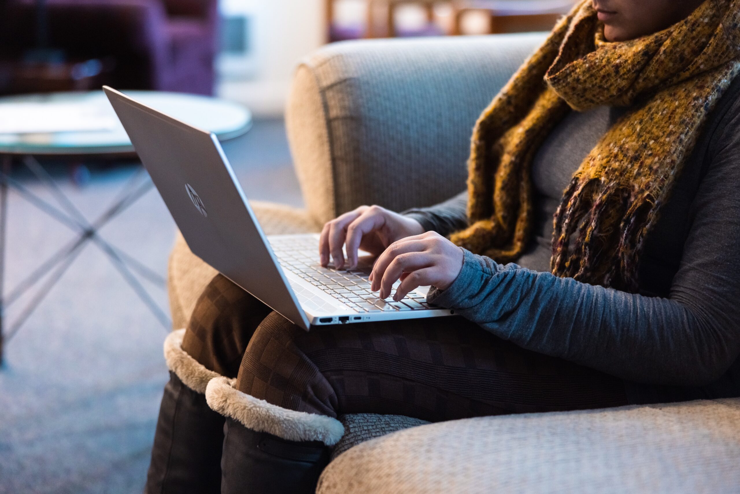 A woman sitting on a couch, focused on her laptop, with a cozy living room setting in the background.
