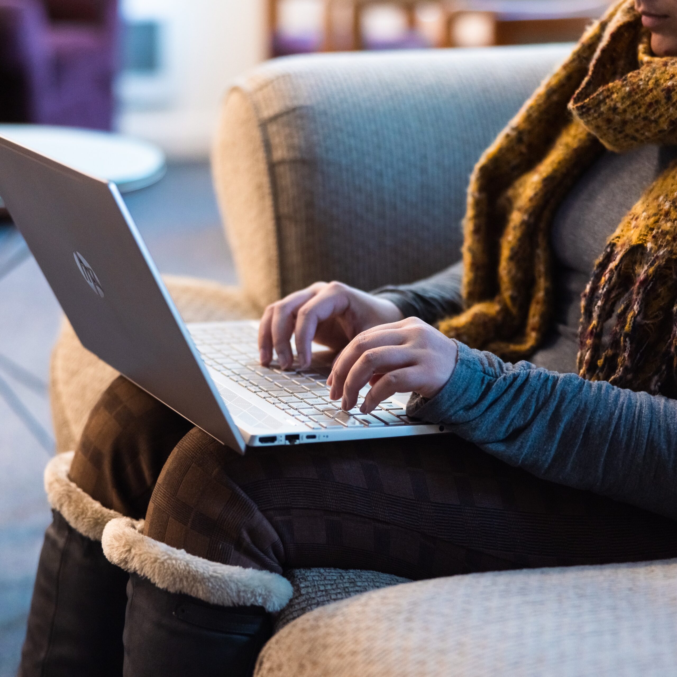 A woman sitting on a couch, focused on her laptop, with a cozy living room setting in the background.
