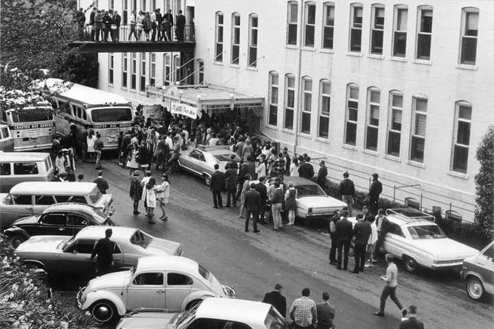 Black and white photograph featuring cars parked in front of the Schimmel building and students milling around, taken in 1970.