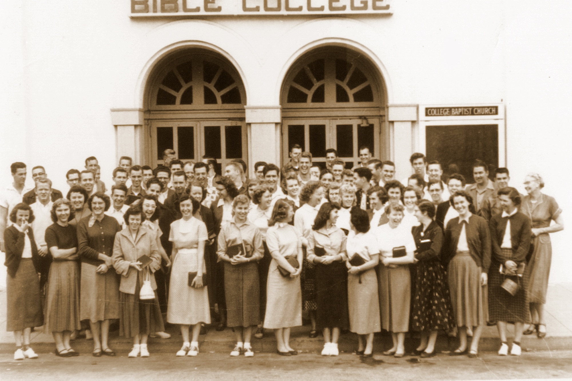 A group of students standing in front of the Oakland campus building, smiling.