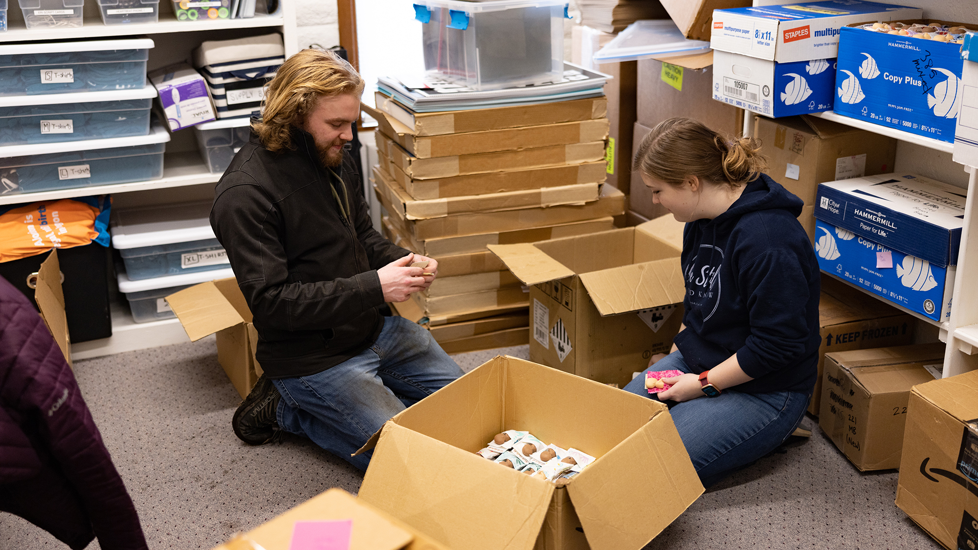 Two people seated on the floor, sorting through boxes for Serve Day in a room filled with supplies.