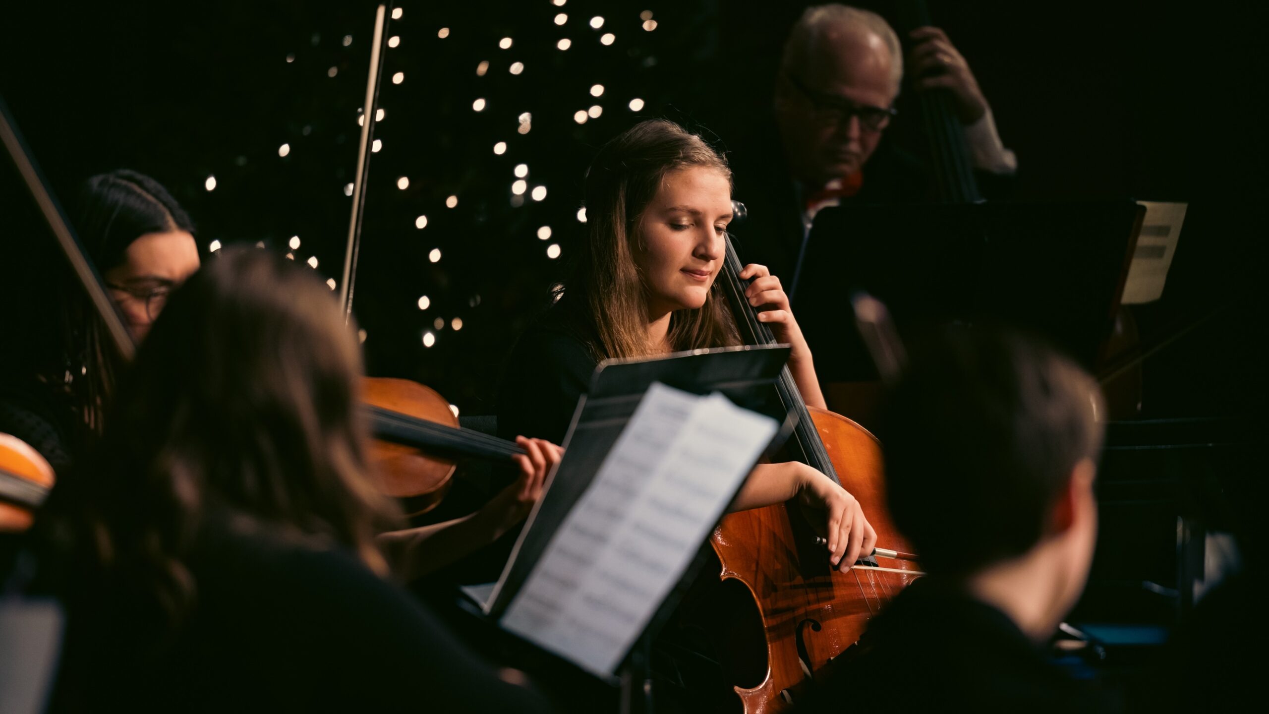 A woman is playing the cello while surrounded by fellow musicians for Christmas at Corban.