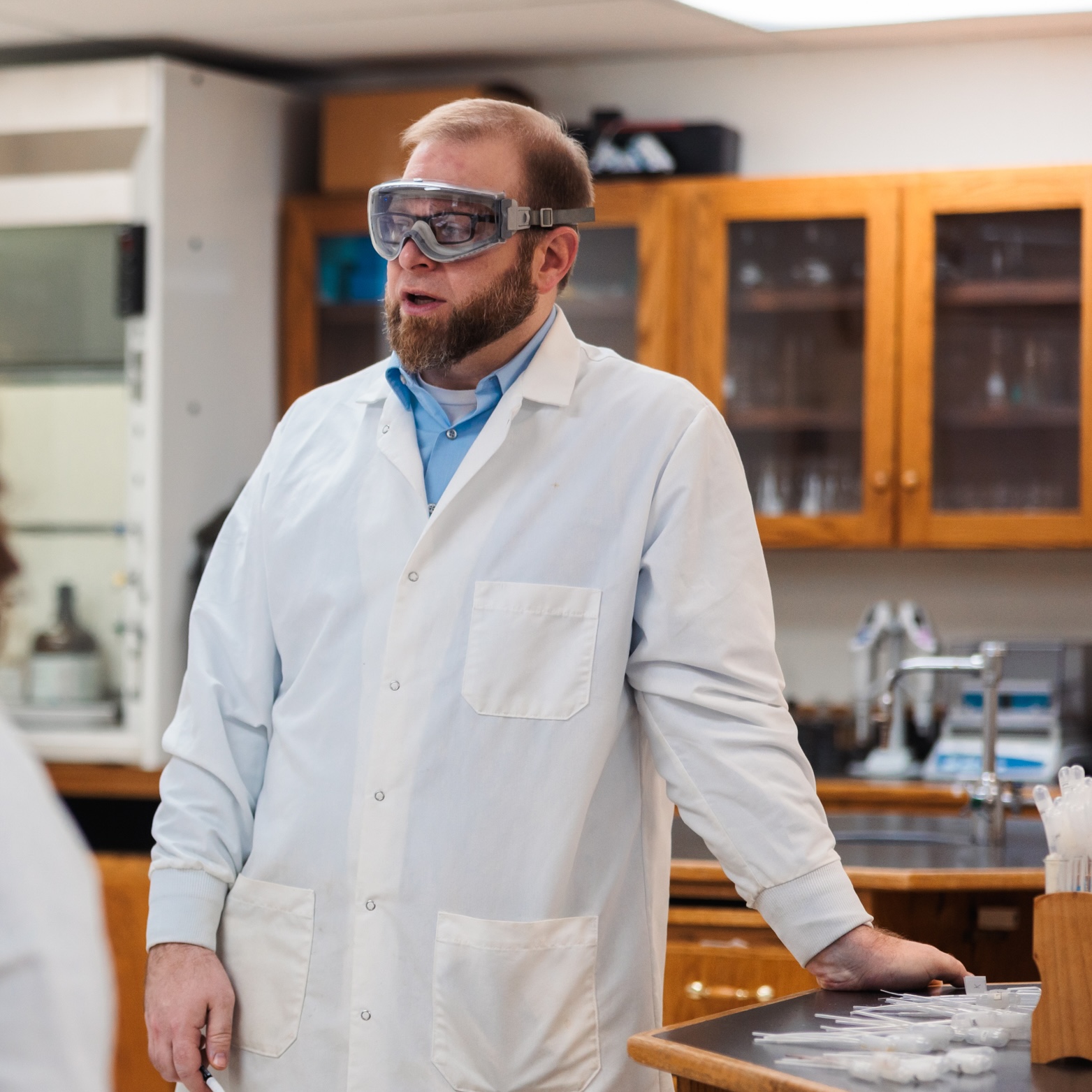 Clinton Knaus wearing a lab coat, standing in a laboratory setting, talking about research.