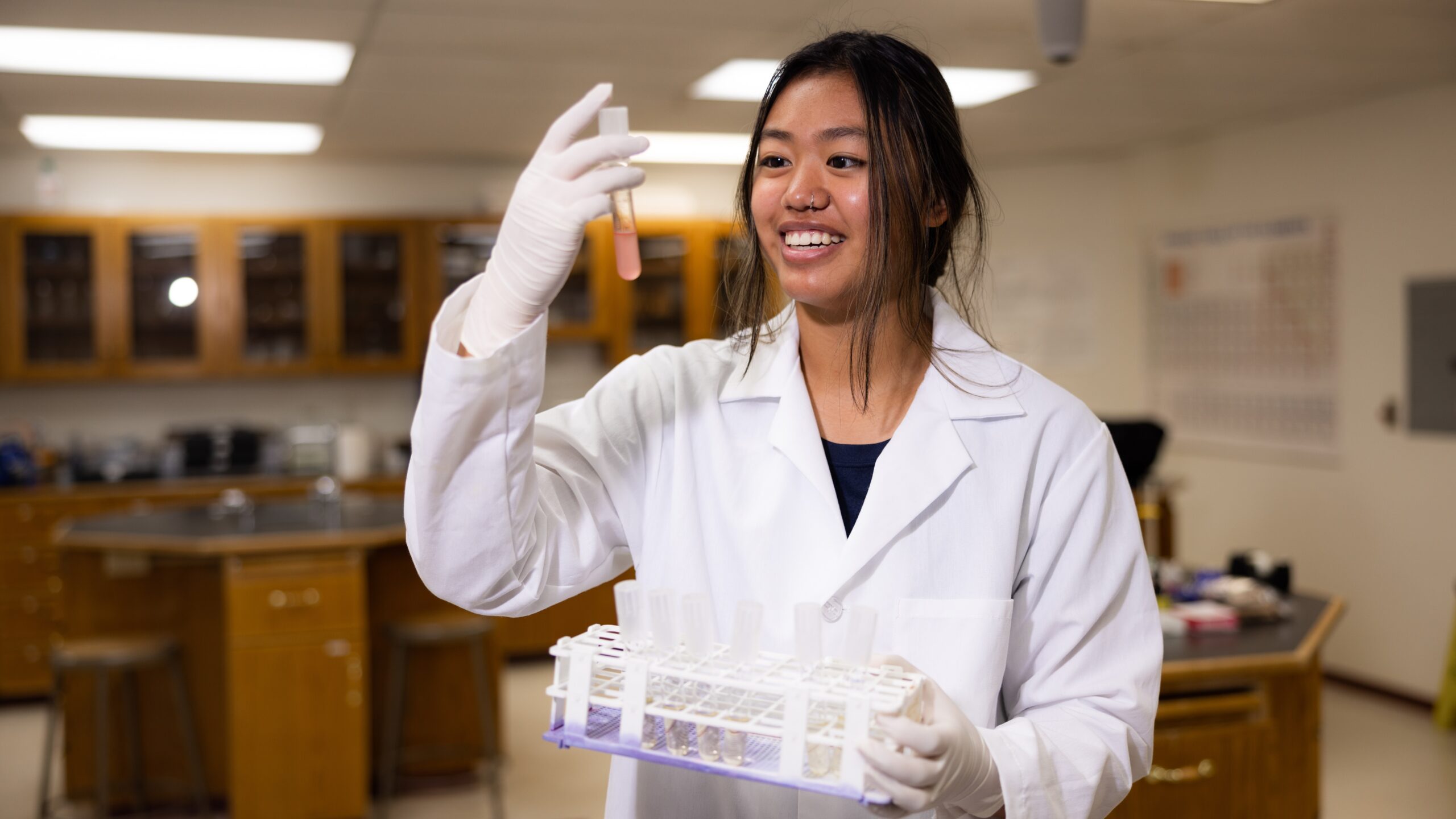 A female scientist in a lab coat holds a test tube filled with a colorful liquid, smiling confidently.