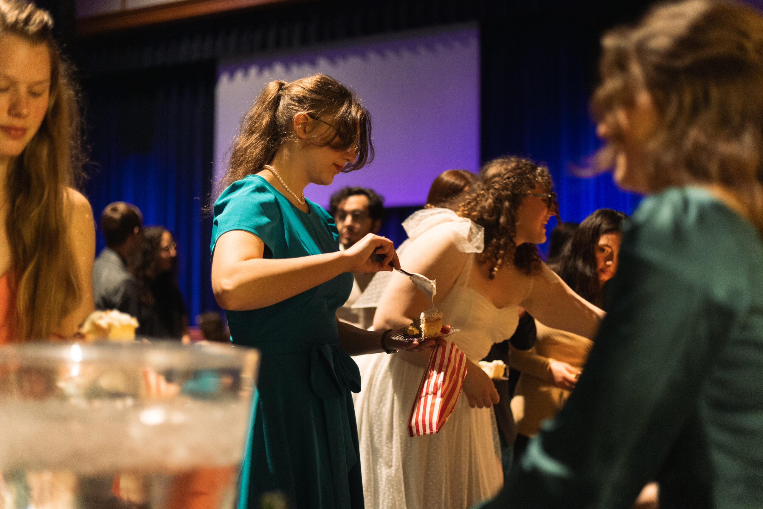 People in formal attire serve themselves dessert from a buffet table at an indoor event, with dim lighting and a blue-lit background.