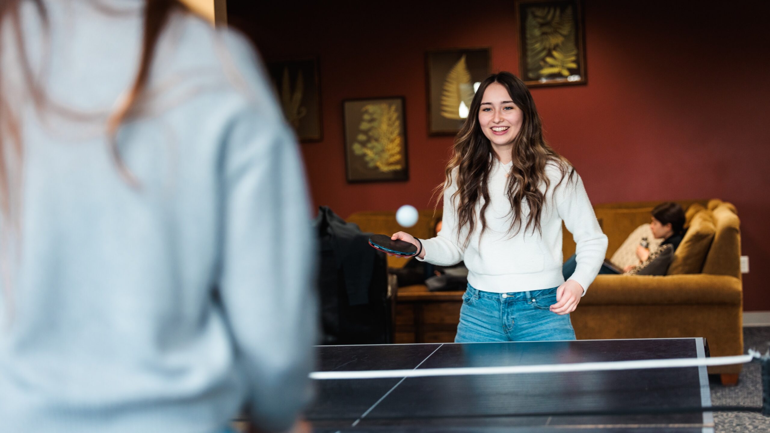 A woman playing ping pong in a dorm lobby, participating in an intramural game.