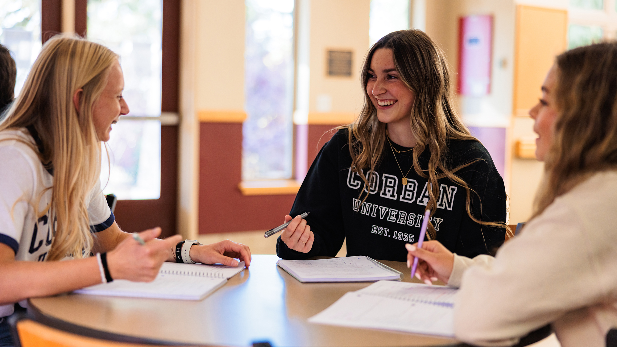 Three female students gather at a table, writing in notebooks and sharing ideas during a study session.