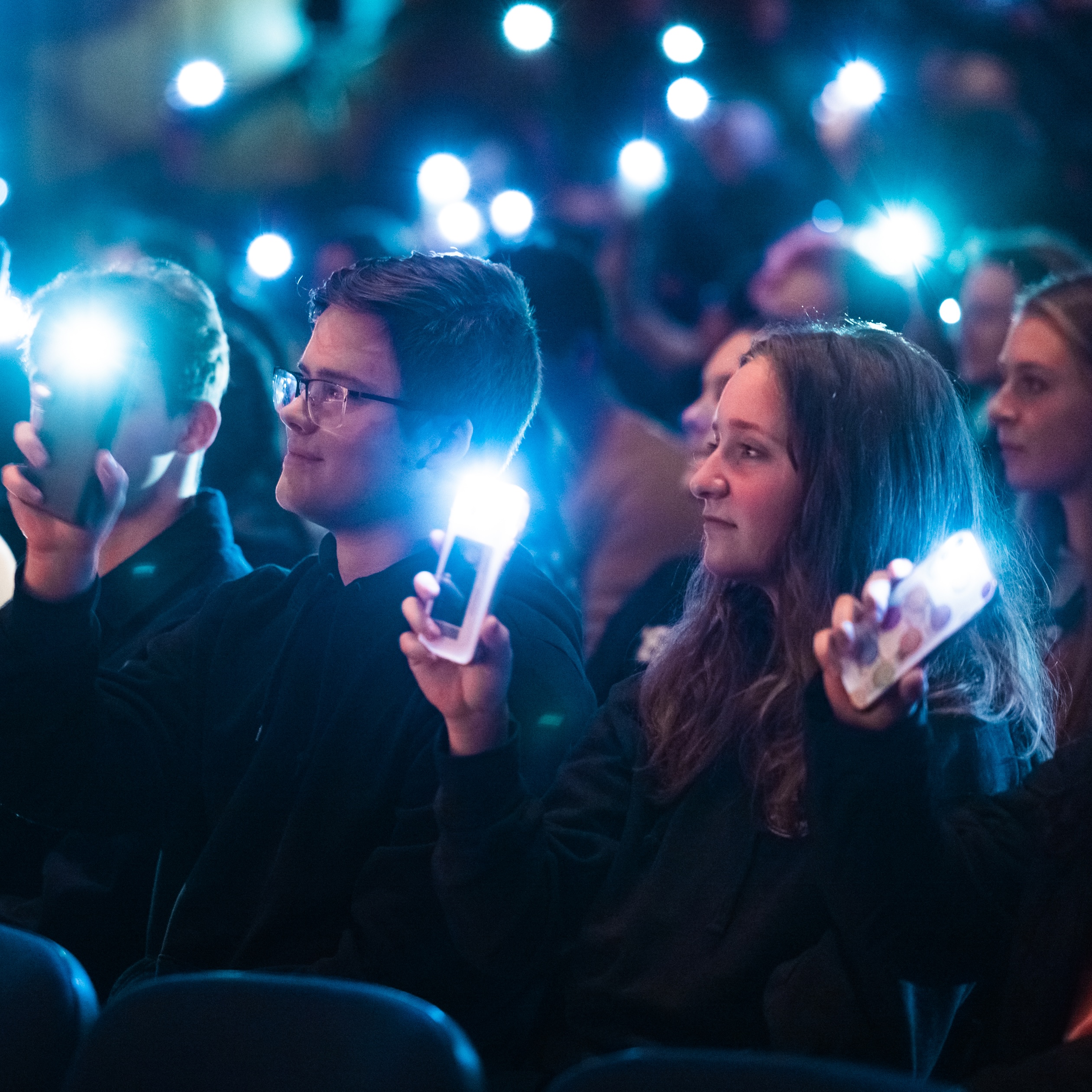 Audience members in a dimly lit auditorium hold up phones with flashlight apps turned on. They appear engaged and focused on an unseen event, with expressions of interest and small smiles visible. The setting suggests a concert or performance.