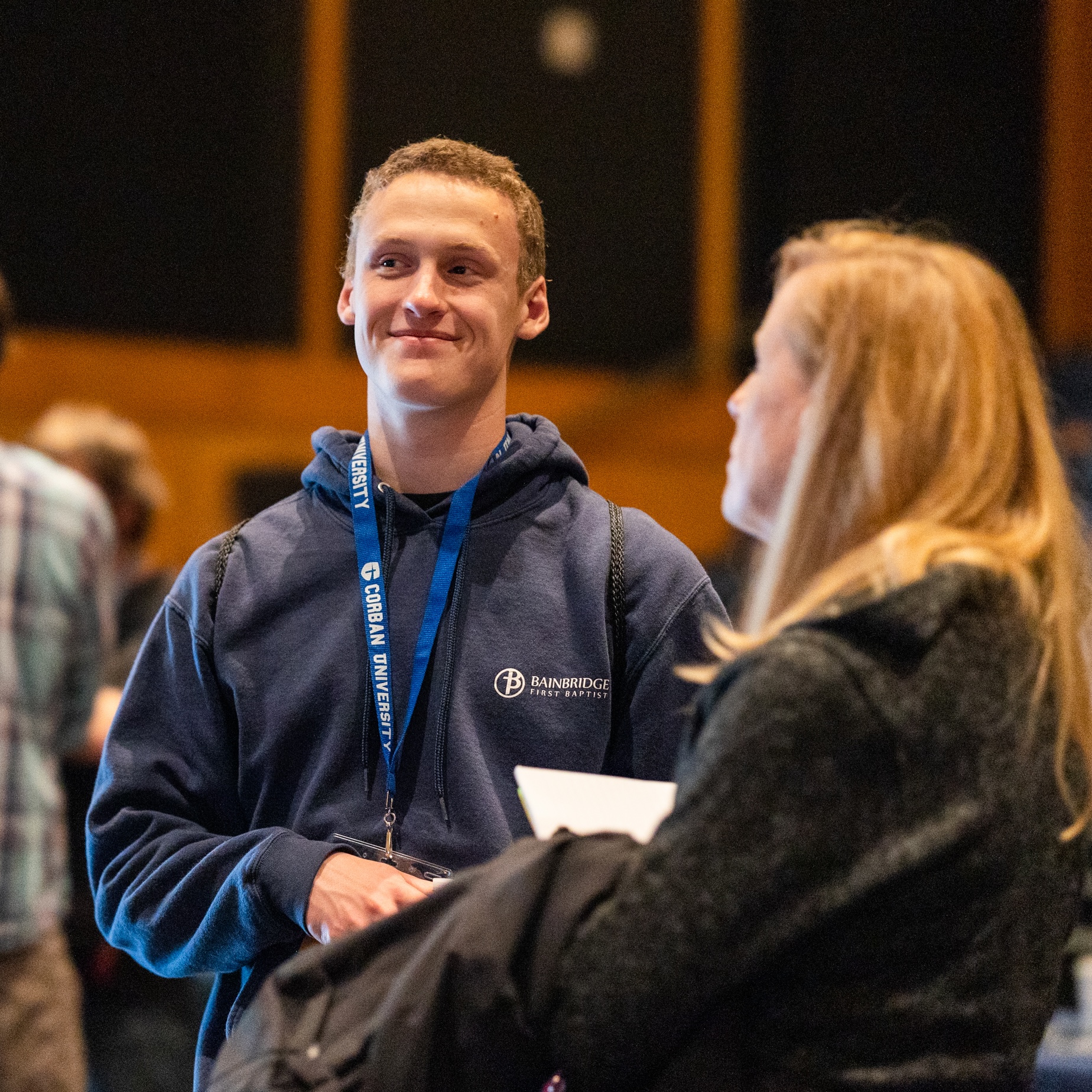 A young man wearing a navy hoodie and a lanyard smiles while engaging in conversation with two people. They are in a warmly lit indoor setting with blurred figures and seating in the background.