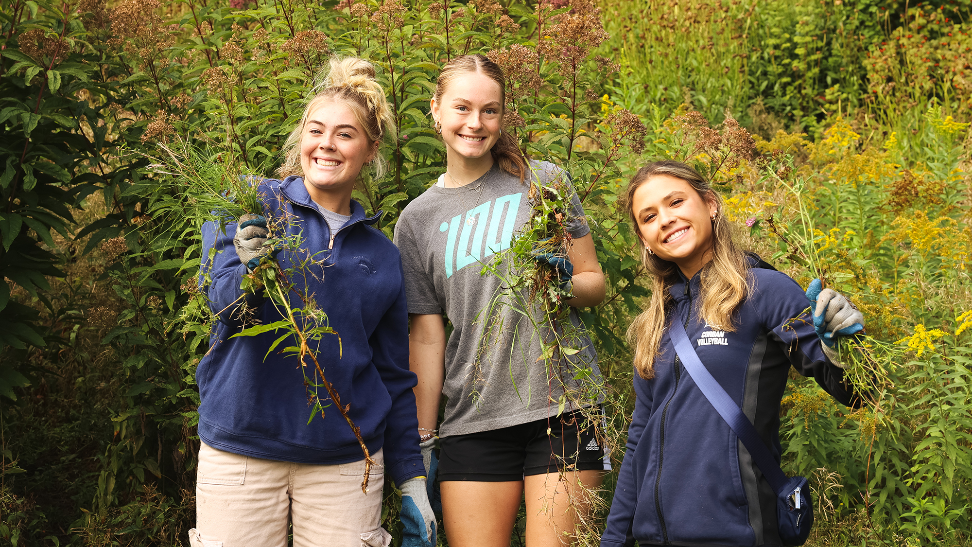 Three girls in blue shirts smiling as they hold weeds they've pulled while serving as volunteers.
