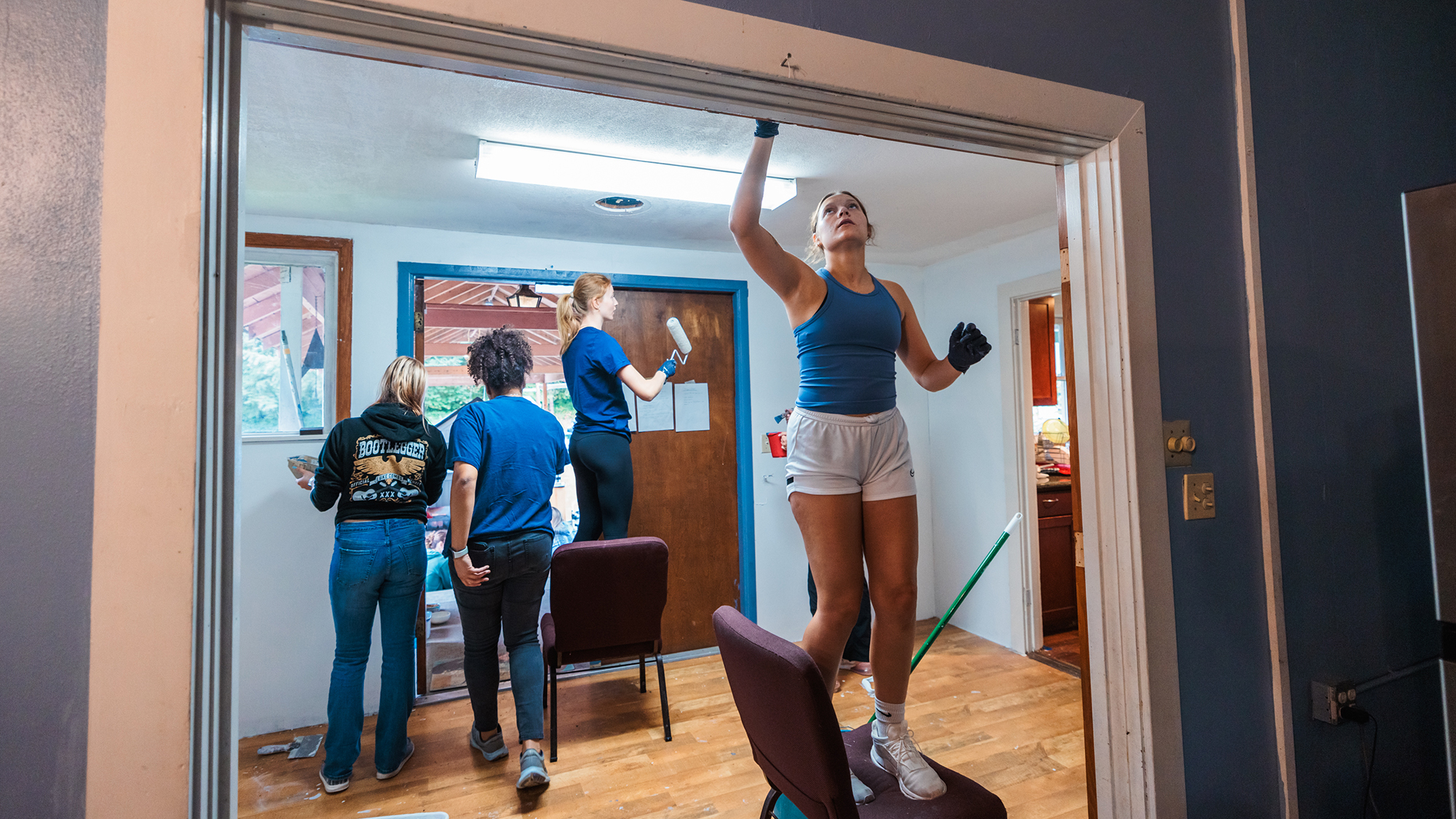 A woman stands on a chair in a doorway, painting trim as a volunteer on Serve Day.