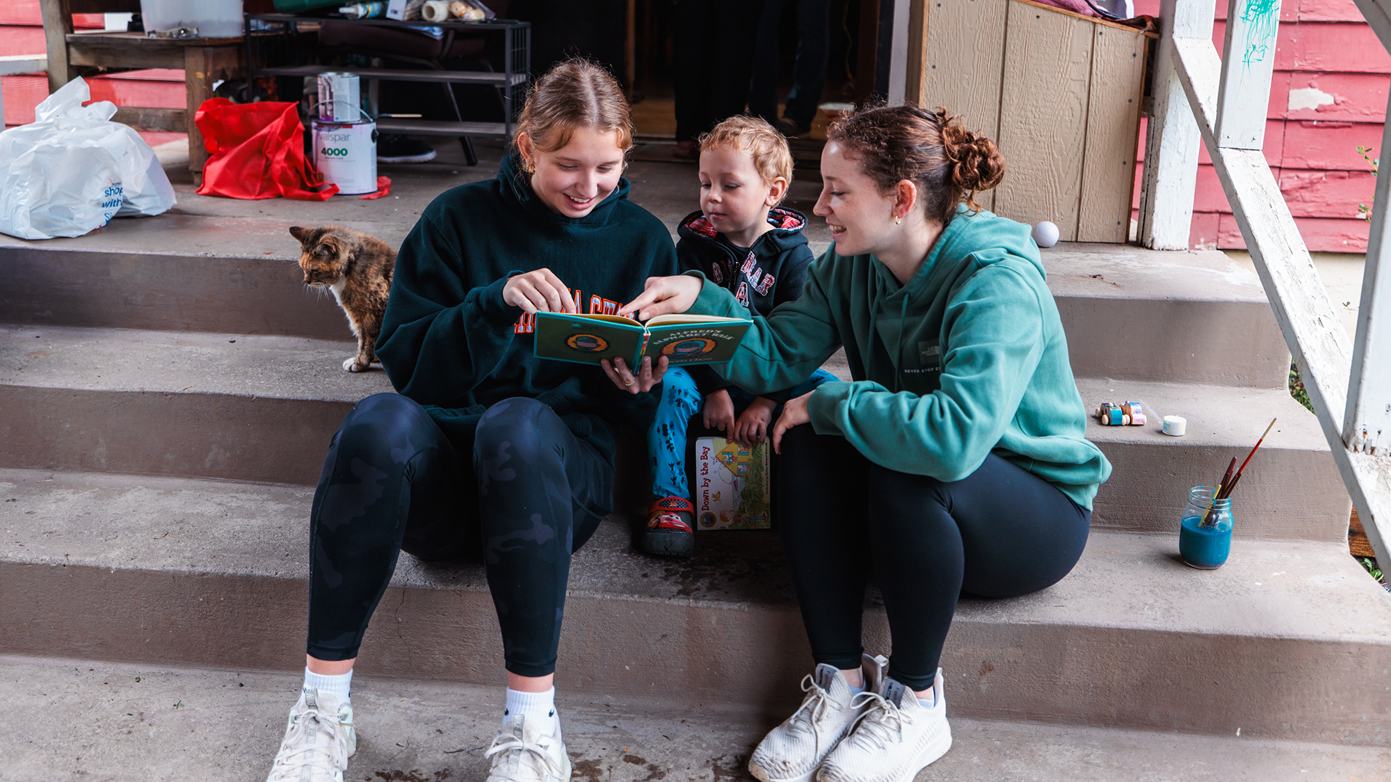 Two women sit on porch steps and read a book to a young child during Serve Day.