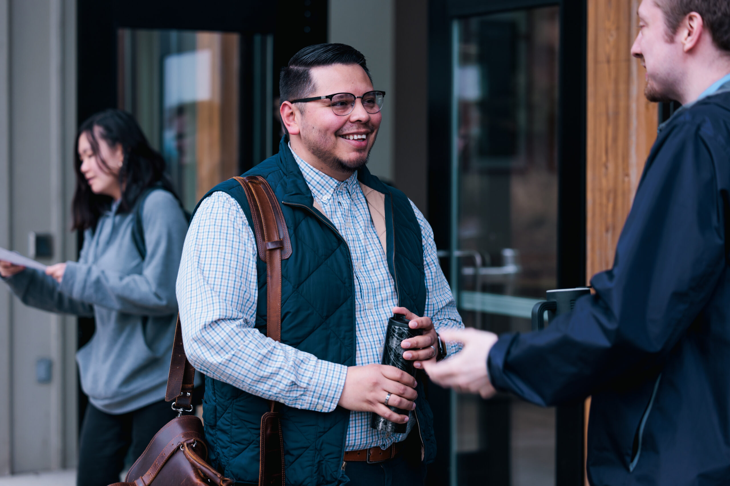 A graduate student carrying a brown leather bag over his shoulder, dressed casually in a light shirt and jeans.