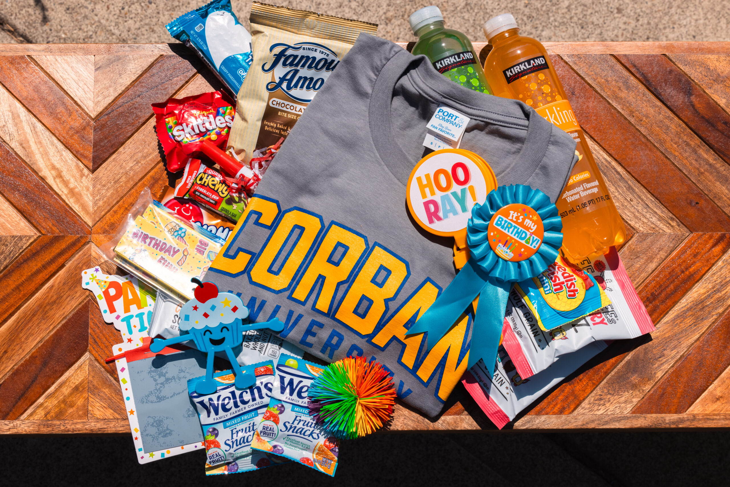 A Corban University t-shirt displayed with snacks and goodies, including Skittles, Cheez-It, Famous Amos cookies, fruit snacks, a pom-pom, two juice bottles, and festive Hooray! ribbons, all arranged on a wooden surface.