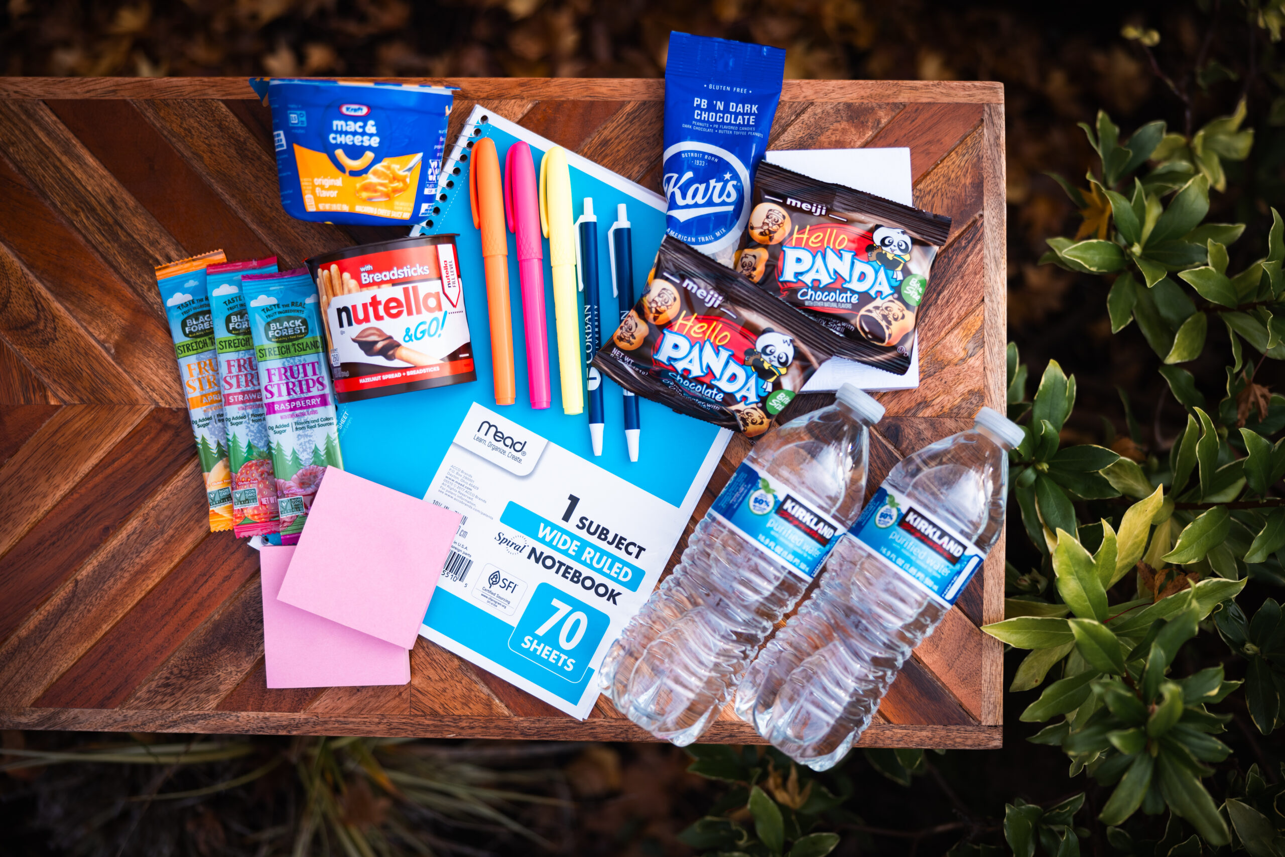 A collection of school supplies and snacks on a wooden table, including a spiral notebook, pens, highlighters, sticky notes, Nutella to-go, Hello Panda cookies, fruit strips, water bottles, mac & cheese, and peanuts.