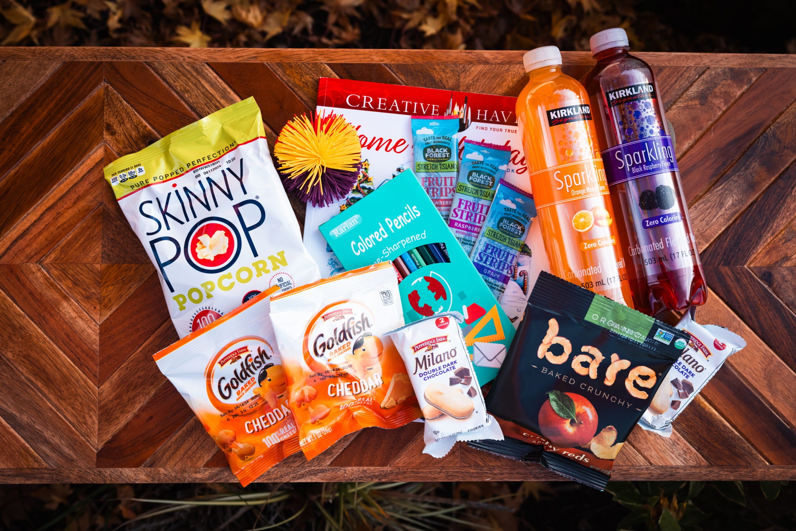 A variety of snacks and drinks are arranged on a wooden surface, including SkinnyPop popcorn, Goldfish cheddar crackers, Milano cookies, baked apple chips, creative coloring books, and Sparkling Ice drinks in orange and berry flavors.