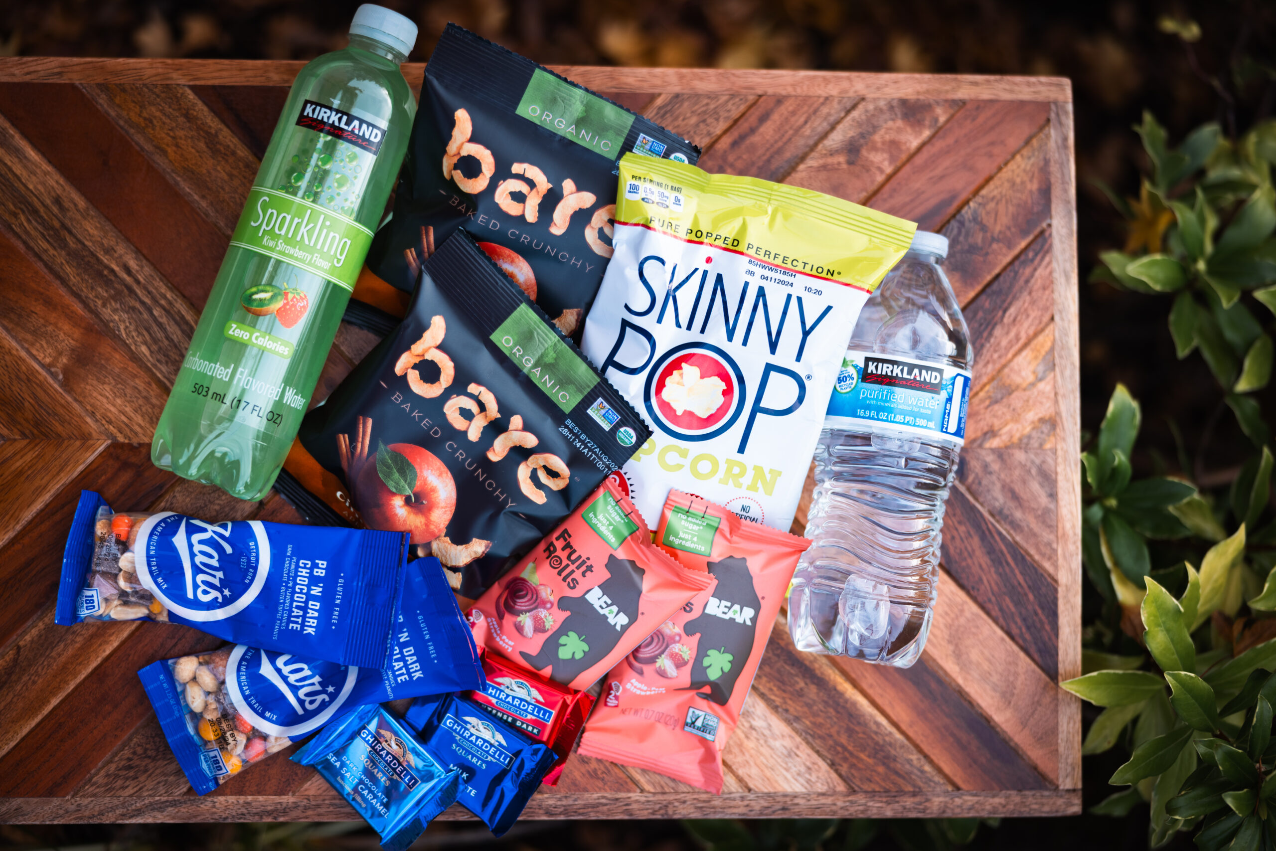 A wooden table displays an assortment of snacks and drinks, including a bottle of sparkling apple cider, bottled water, Bare apple chips, SkinnyPop popcorn, Kind bars, and mini Ritter Sport chocolates.